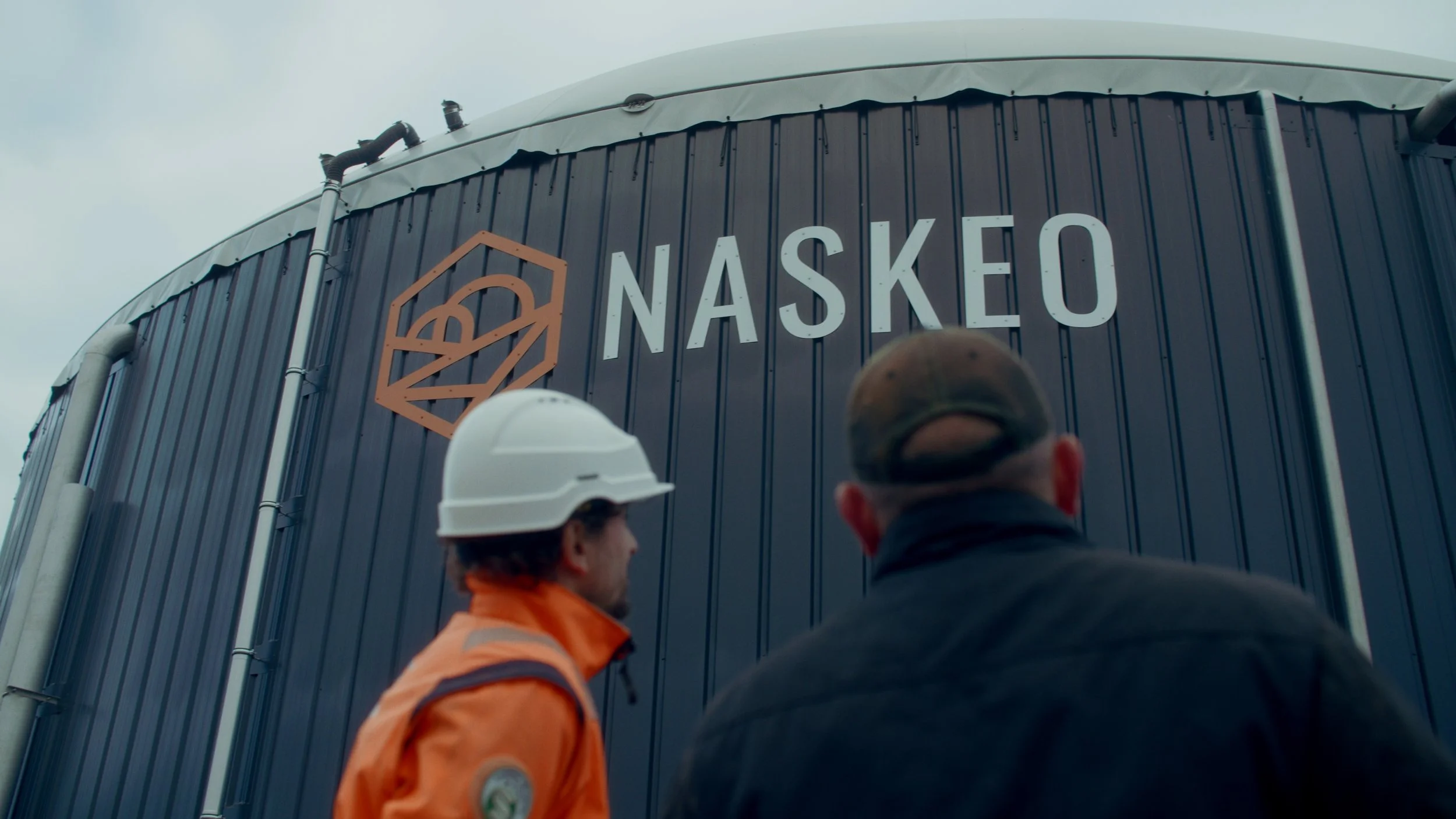 Deux hommes portent des casques de sécurité et regardent un bâtiment avec le logo NASKO écrit dessus.