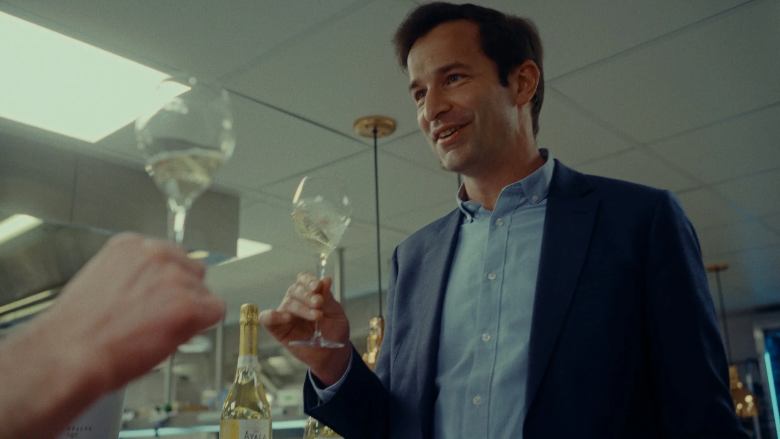 Un homme en costume souriant, tenant deux flûtes à champagne, dans un environnement d'hôtel ou restaurant.