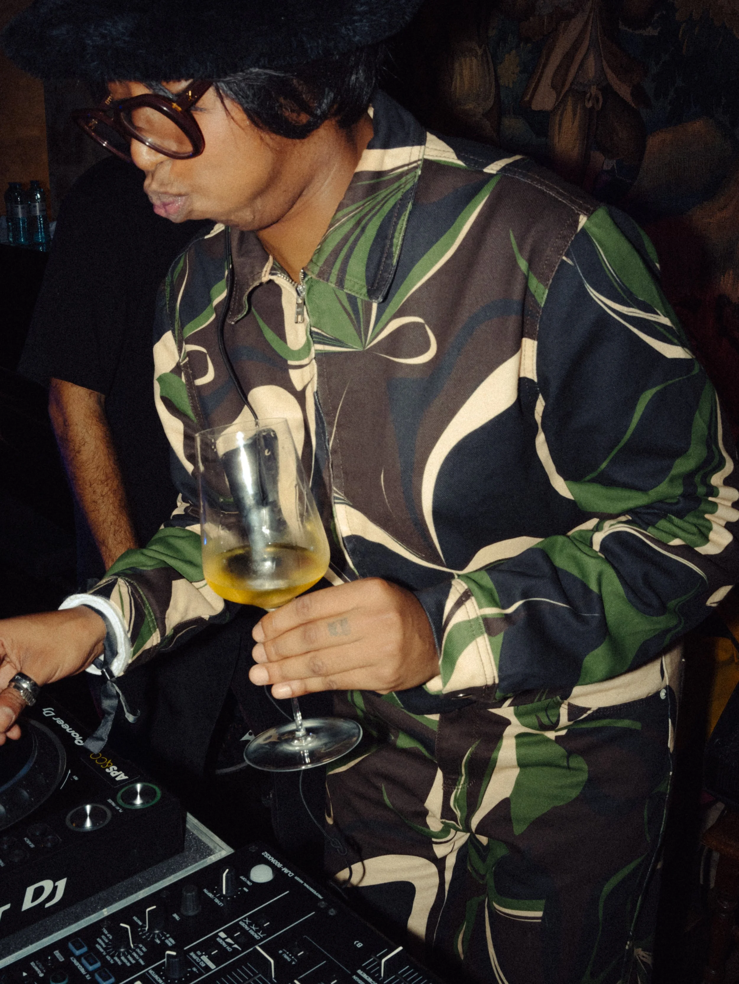Une personne portant des lunettes épaisses et un vêtement à motif coloré, tenant un verre de vin blanc, prête à mixer dans un environnement où il y a un DJ.