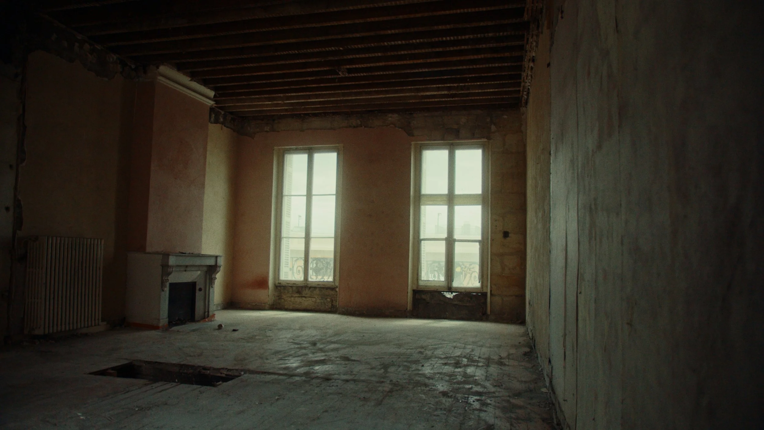 Intérieur d'une pièce en rénovation avec deux grandes fenêtres, un cheminée ancienne, murs en cours de rénovation, plafond en bois apparent, sol en travaux.