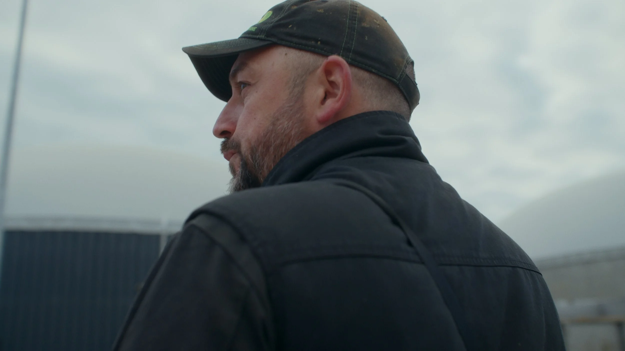 Un homme portant une casquette noire et un manteau noir regarde vers la gauche, avec un ciel couvert en arrière-plan.