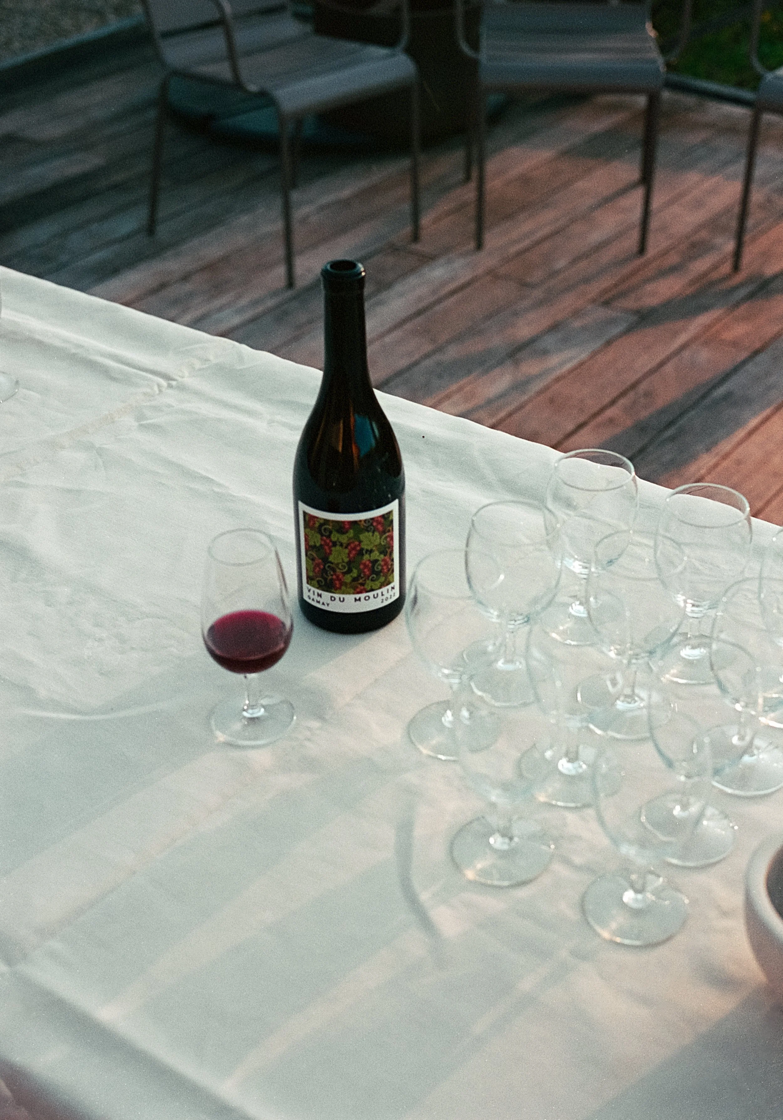 Bouteille de vin et verre de vin rouge sur une table avec plusieurs verres vides, en extérieur avec un plancher en bois et des chaises en arrière-plan.