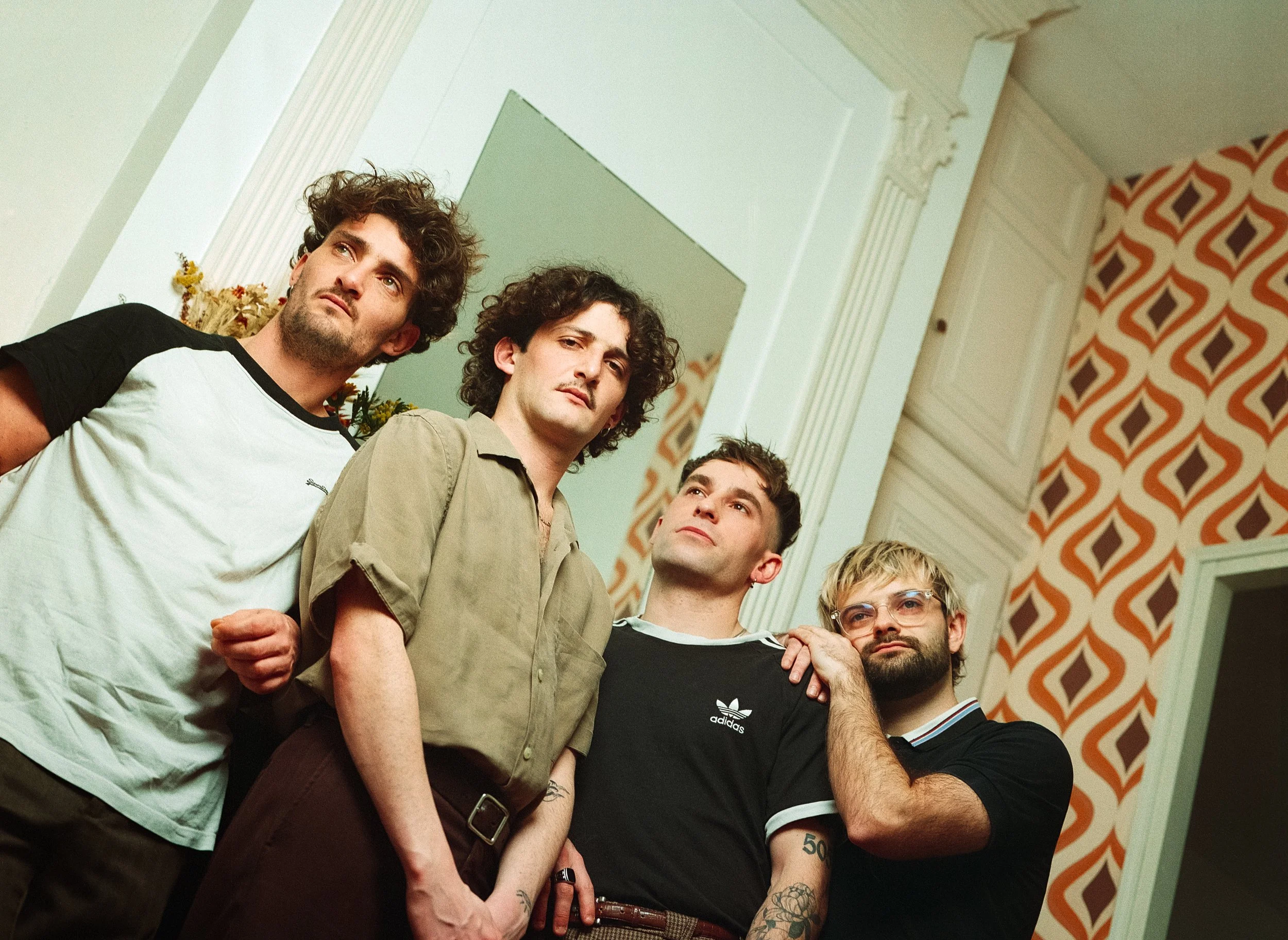 Quatre hommes posant ensemble dans une pièce avec un décor rétro, notamment un mur à motifs oranges et bruns, et un grand miroir dans la pièce.