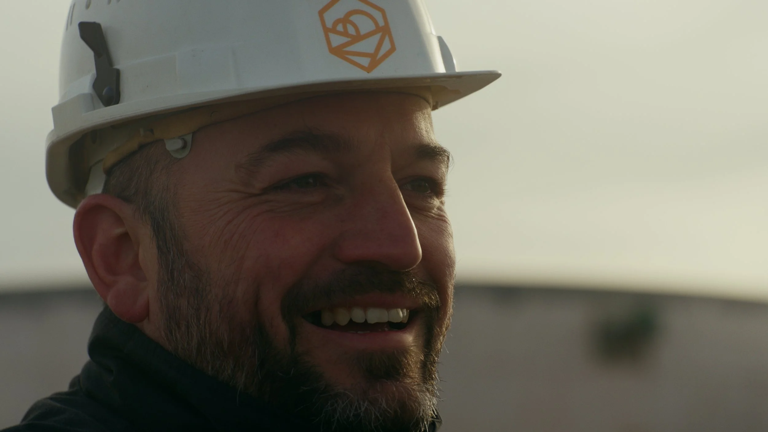 Un homme souriant portant un casque de construction blanc avec un logo orange, en extérieur, sous un ciel clair.