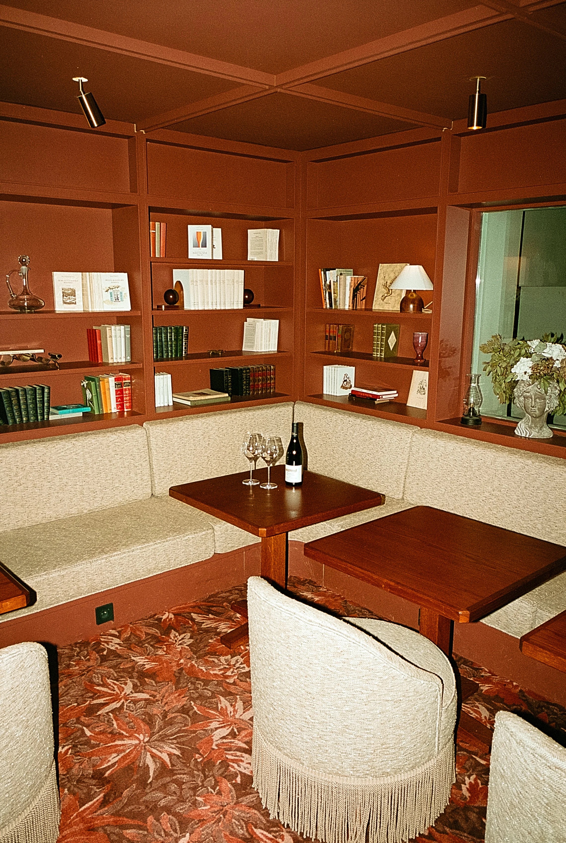 Salon avec des étagères en bois rouge, un canapé en tissu beige, une table en bois avec une bouteille de vin et des verres, un fauteuil beige avec des franges, et un tapis à motif floral rouge et beige.