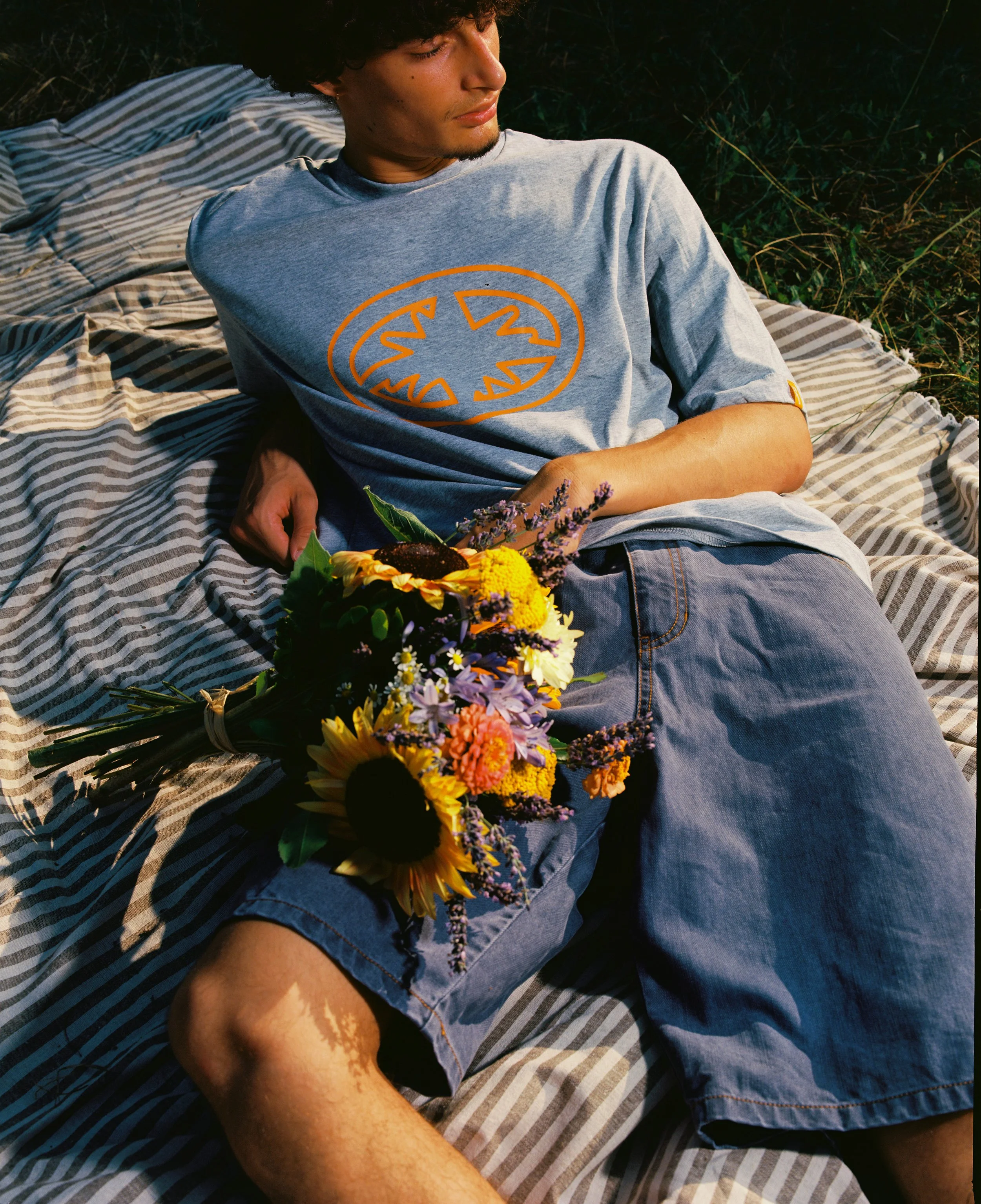 Un jeune homme allongé sur une couverture rayée, tenant un bouquet de fleurs diverses, portant un t-shirt gris avec un logo orange et un short en denim.