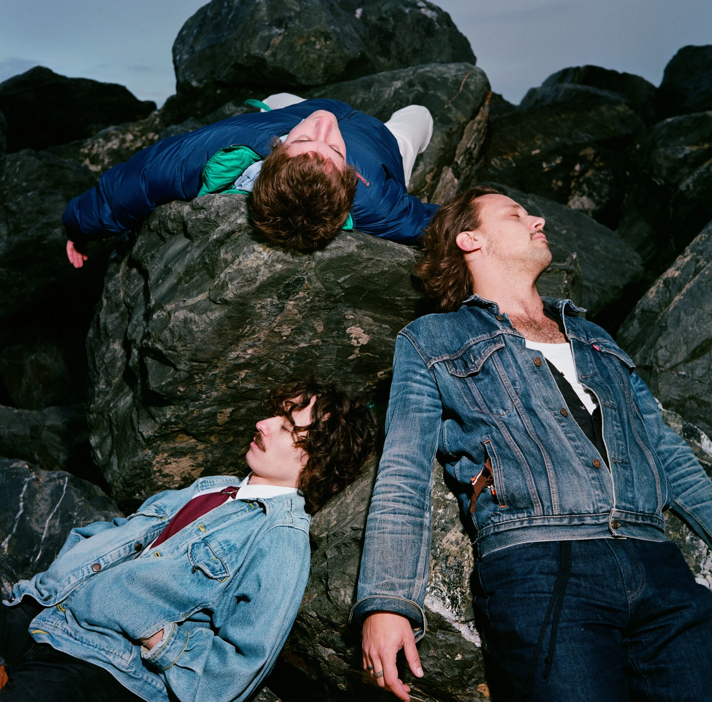 Quatre jeunes hommes dorment sur des rochers, certains allongés et d'autres inclinés, en plein air, probablement près de la plage ou d'un bord de mer.