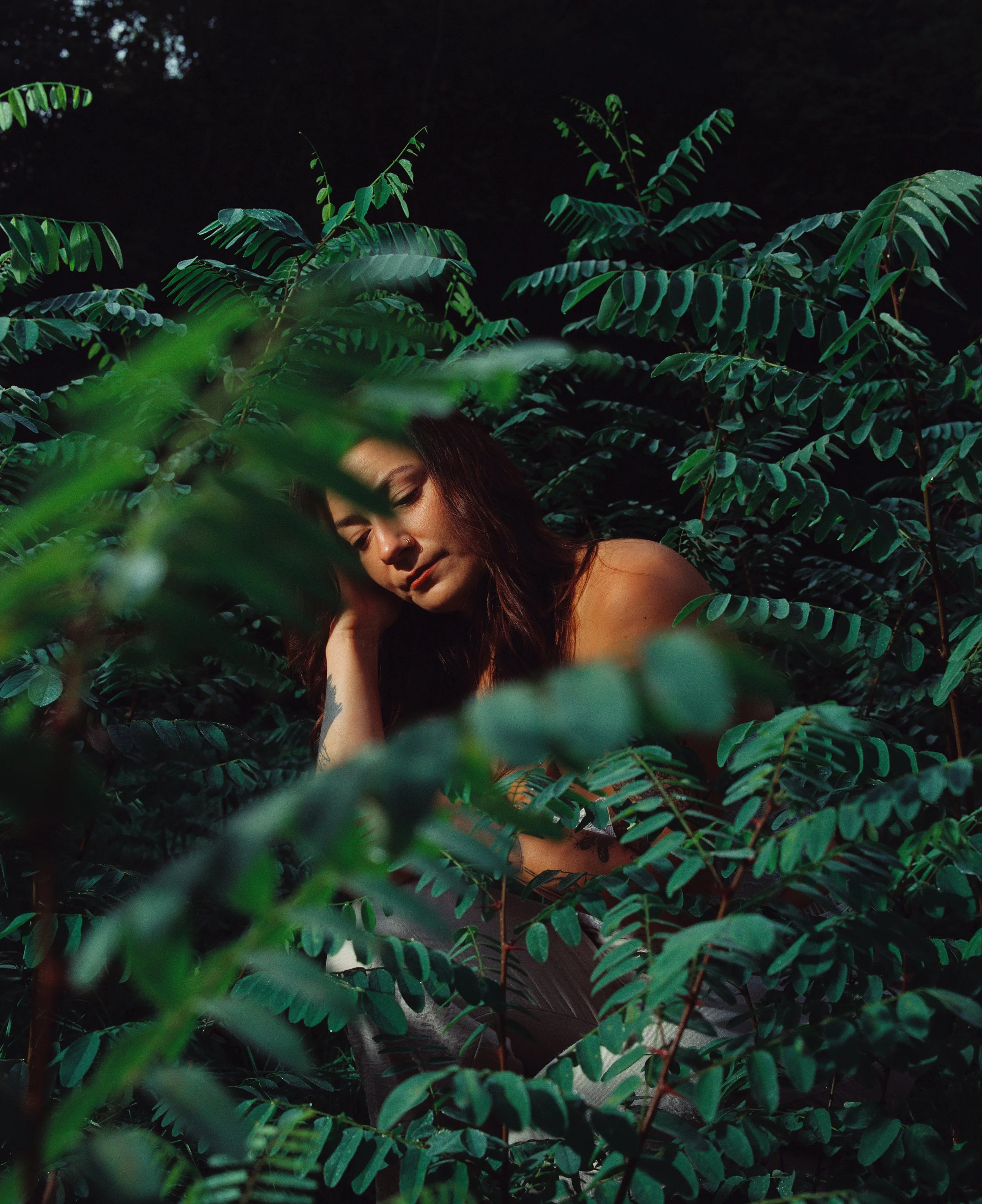 Une femme aux cheveux bruns, à peau foncée, repose dans la végétation luxuriante d'une forêt ou d'une jungle, avec une expression calme et paisible.