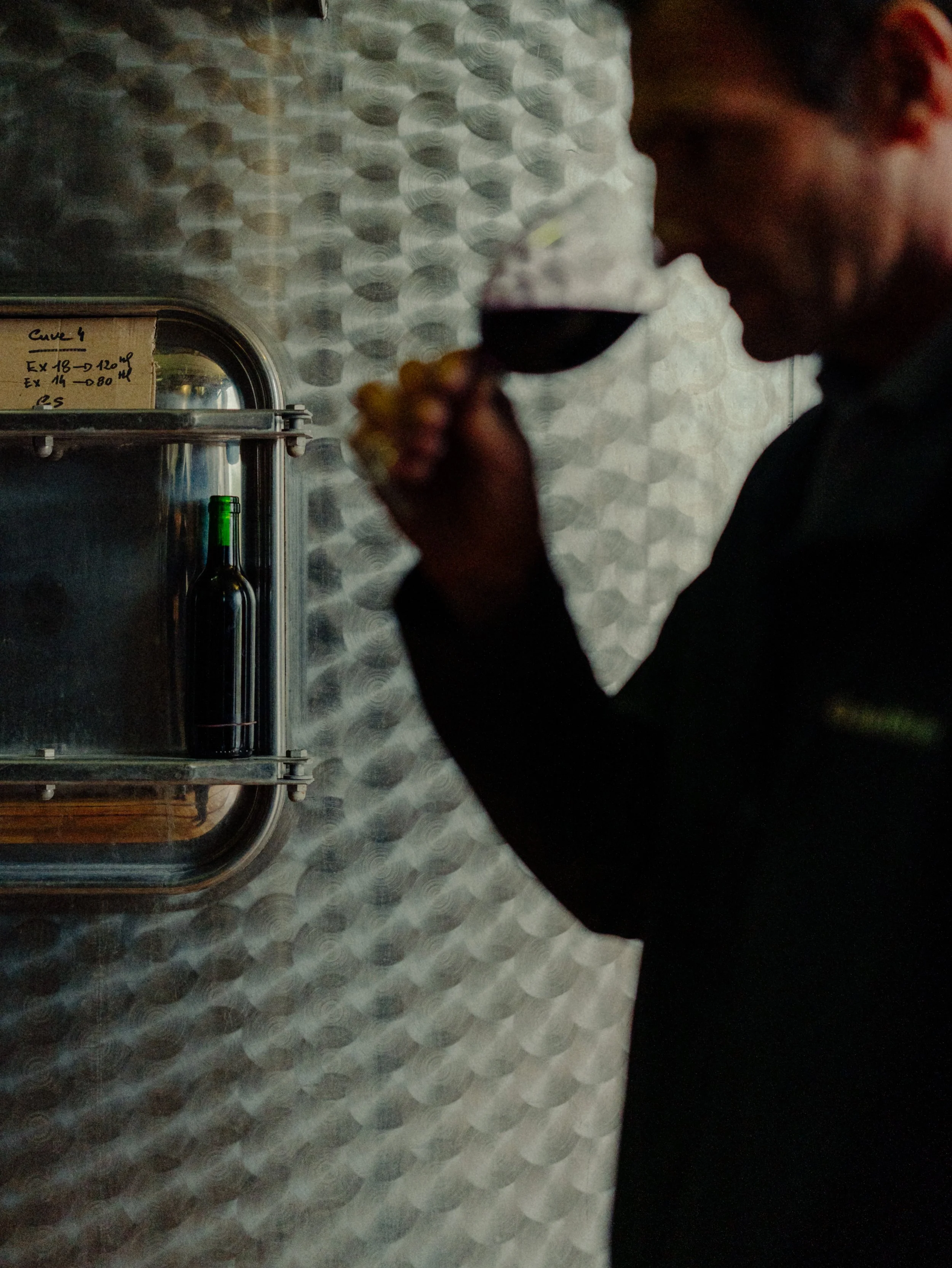 Homme dégustant un verre de vin rouge dans un espace de dégustation ou de cave à vin, avec une bouteille de vin à l'arrière-plan et un mur en métal bRound.