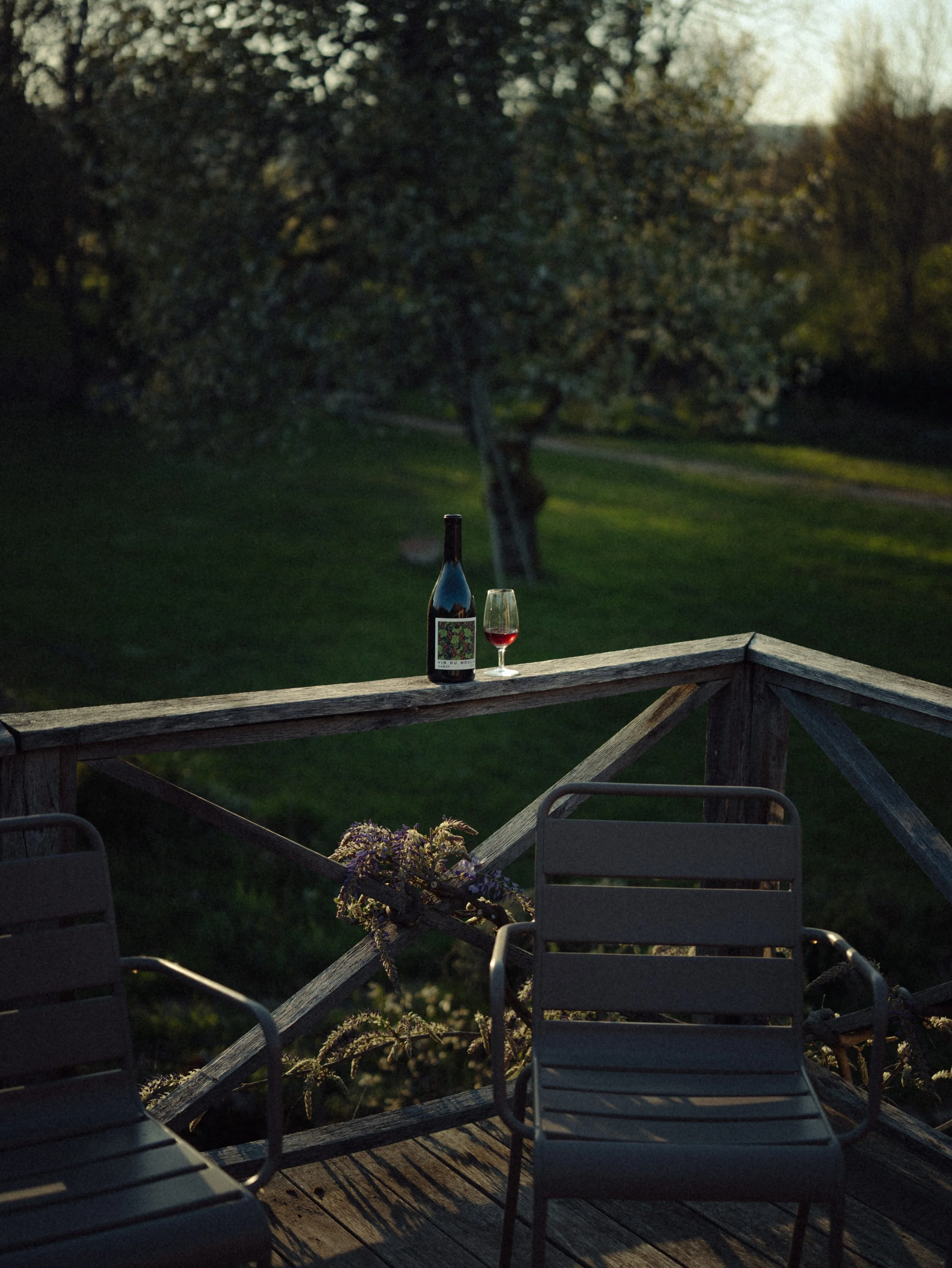 Une bouteille de vin rouge et un verre de vin rouge sur une rambarde en bois d'une terrasse avec deux chaises, au crépuscule dans un environnement extérieur verdoyant.