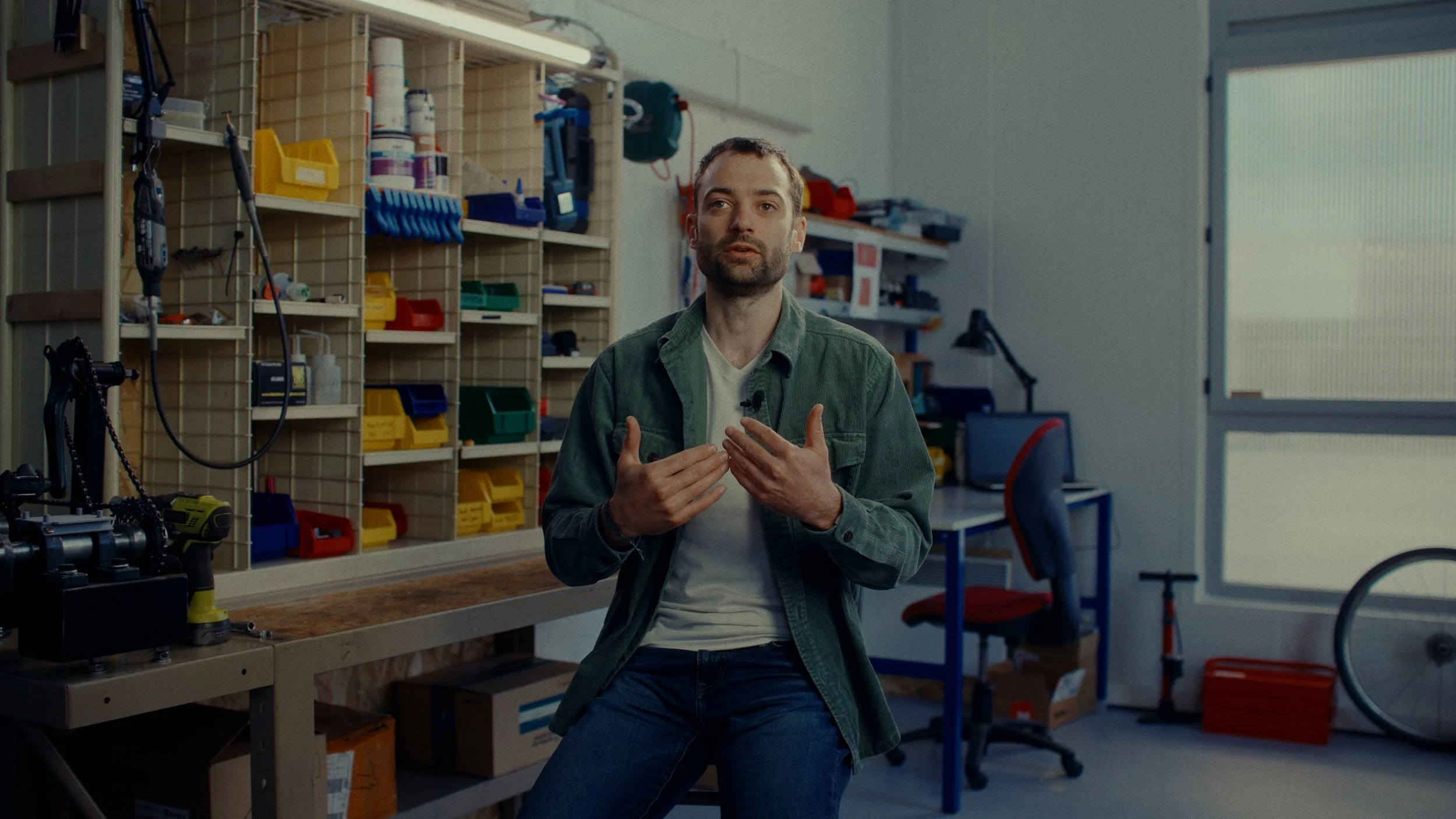 Un homme est assis dans un atelier ou un espace de bricolage, parlant ou donnant une présentation avec des mains. En arrière-plan, il y a des étagères avec des boîtes de rangement colorées, des outils, une table de travail, et un bureau avec une chai