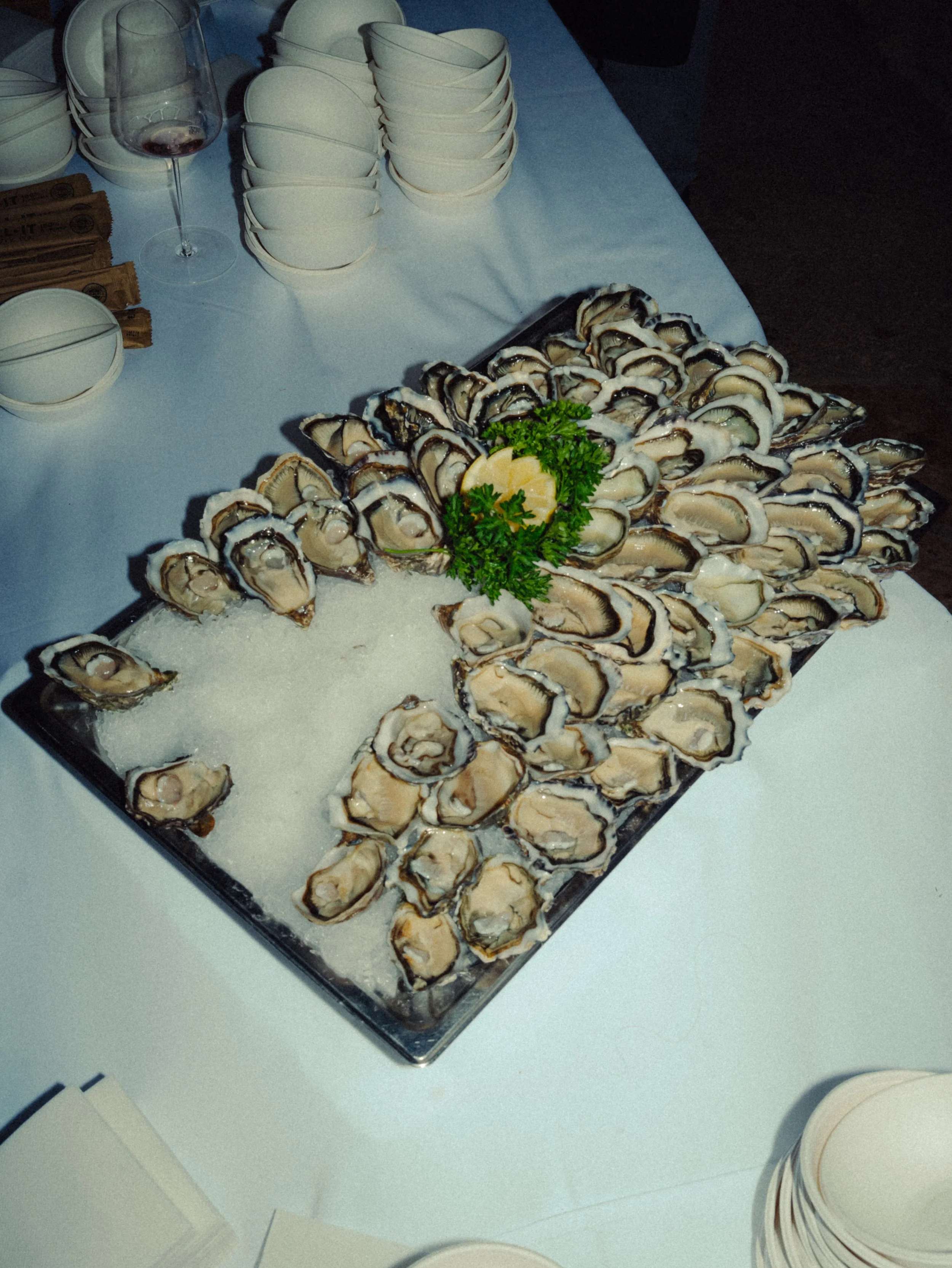 Plate d'huîtres fraîches décorée de persil et de citron, sur un lit de glace, avec des verres de vin blanc et des bols de condensation à proximité.