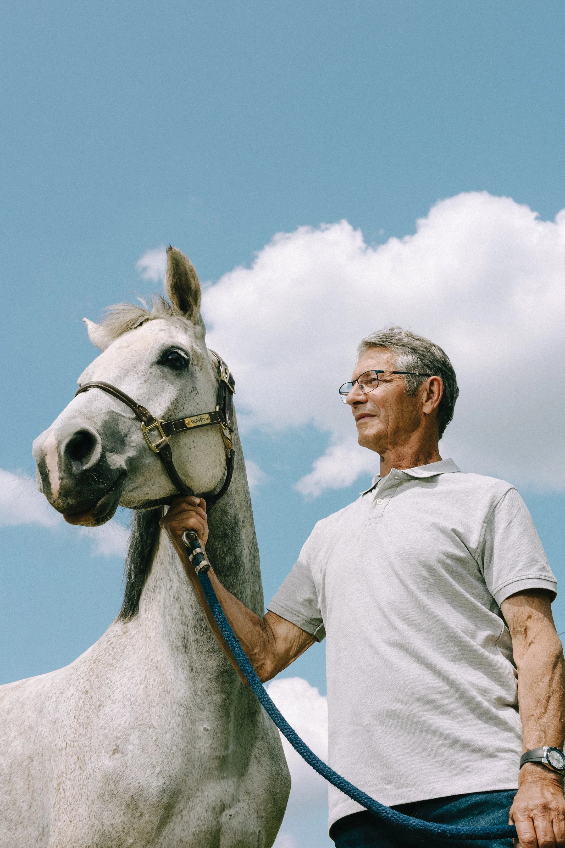 Un homme âgé avec des lunettes tient la bride d'un cheval blanc dans un ciel bleu avec des nuages.