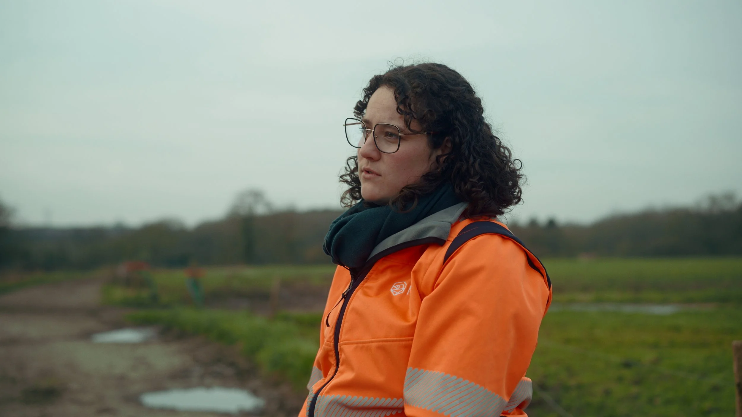 Femme avec lunettes, veste orange, et foulard, debout dans un champ, regardant au loin, temps couvert.
