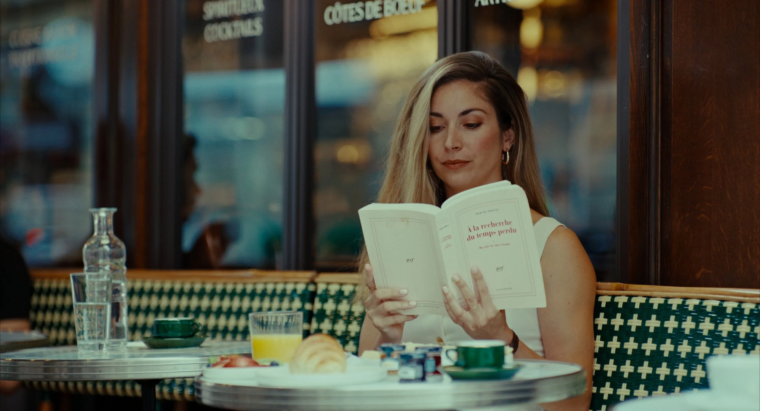 Une femme blonde en robe blanche assise à une table de café, lisant un livre intitulé 'À la recherche du temps perdu'. La table a un verre de jus d'orange, une tasse, une bouteille d'eau, un croissant et des petits pots de confiture.
