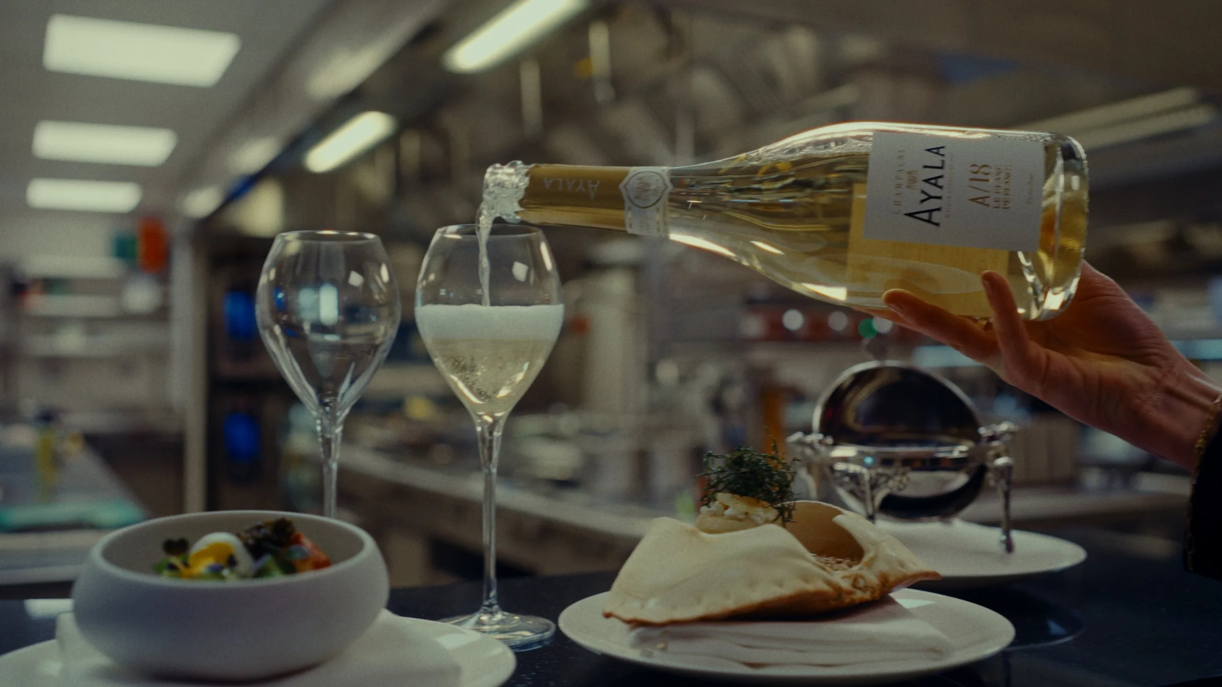 Une main verse du vin blanc dans un verre, avec deux autres verres et une assiette de nourriture dans une cuisine professionnelle.