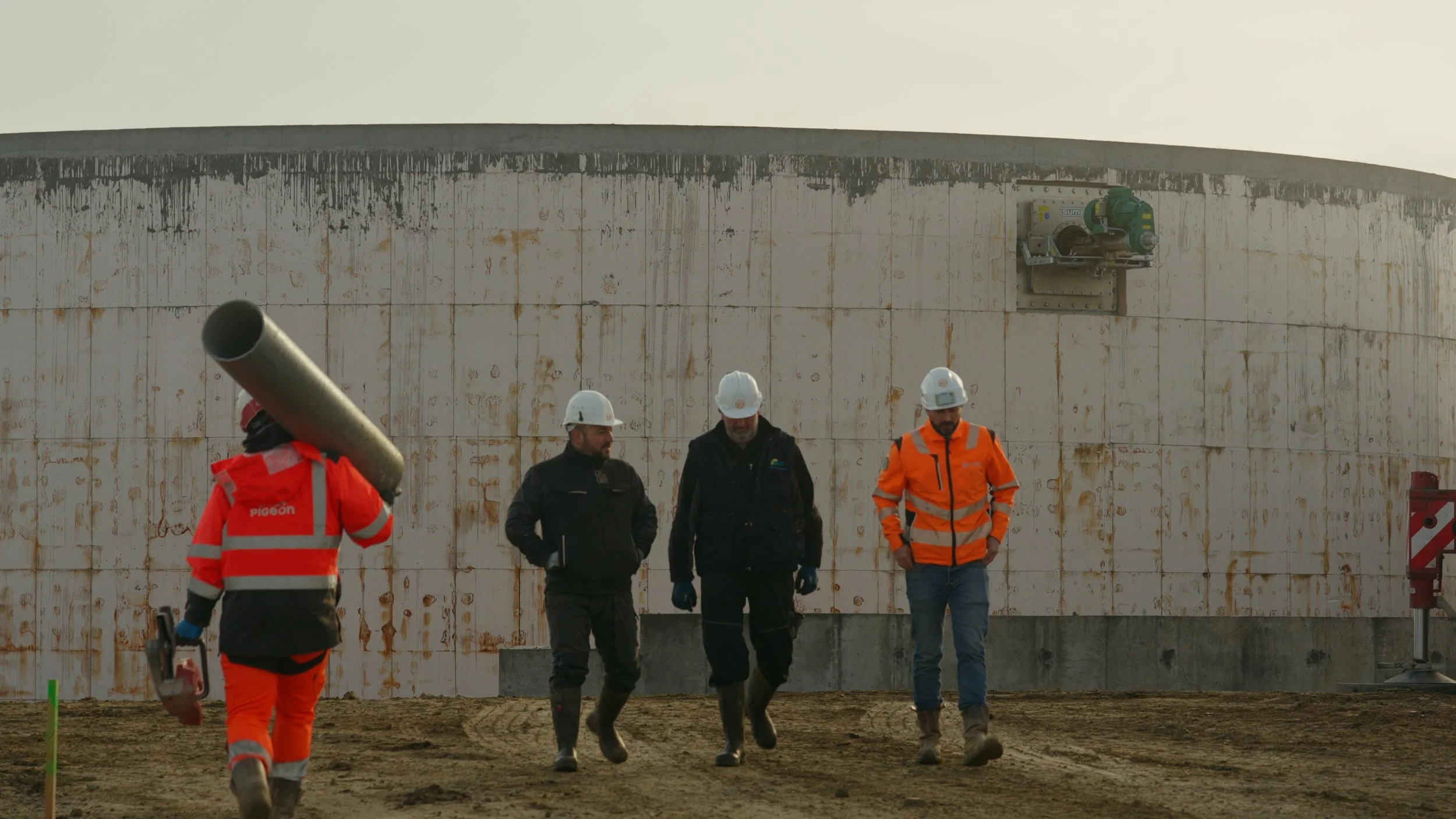 Quatre ouvriers en casque de sécurité et vestes de haute visibilité marchent sur un terrain de construction, avec un grand mur en arrière-plan.