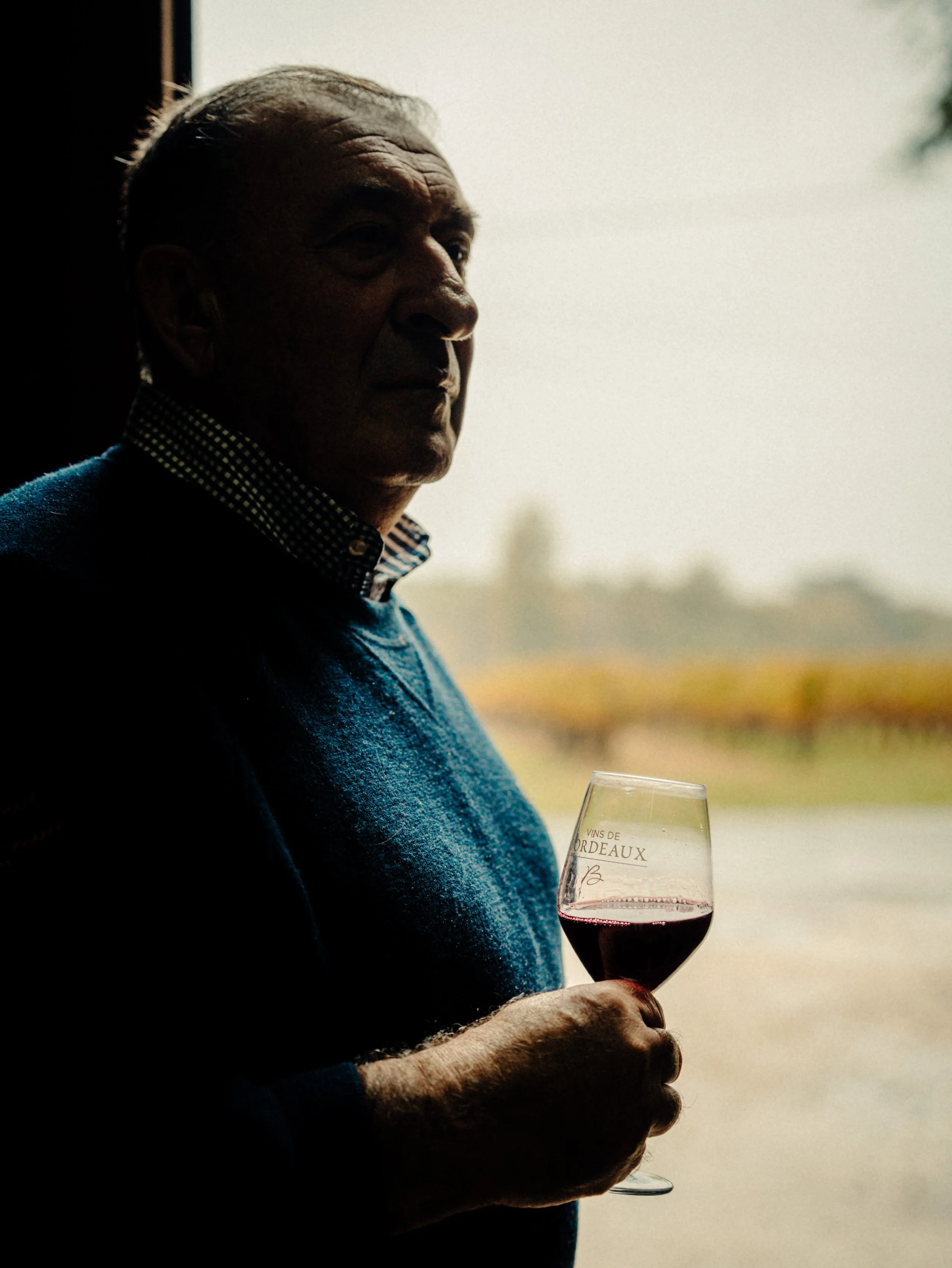 Un homme âgé regarde par une fenêtre en tenant un verre de vin rouge, en arrière-plan un paysage flou avec des champs et des arbres.