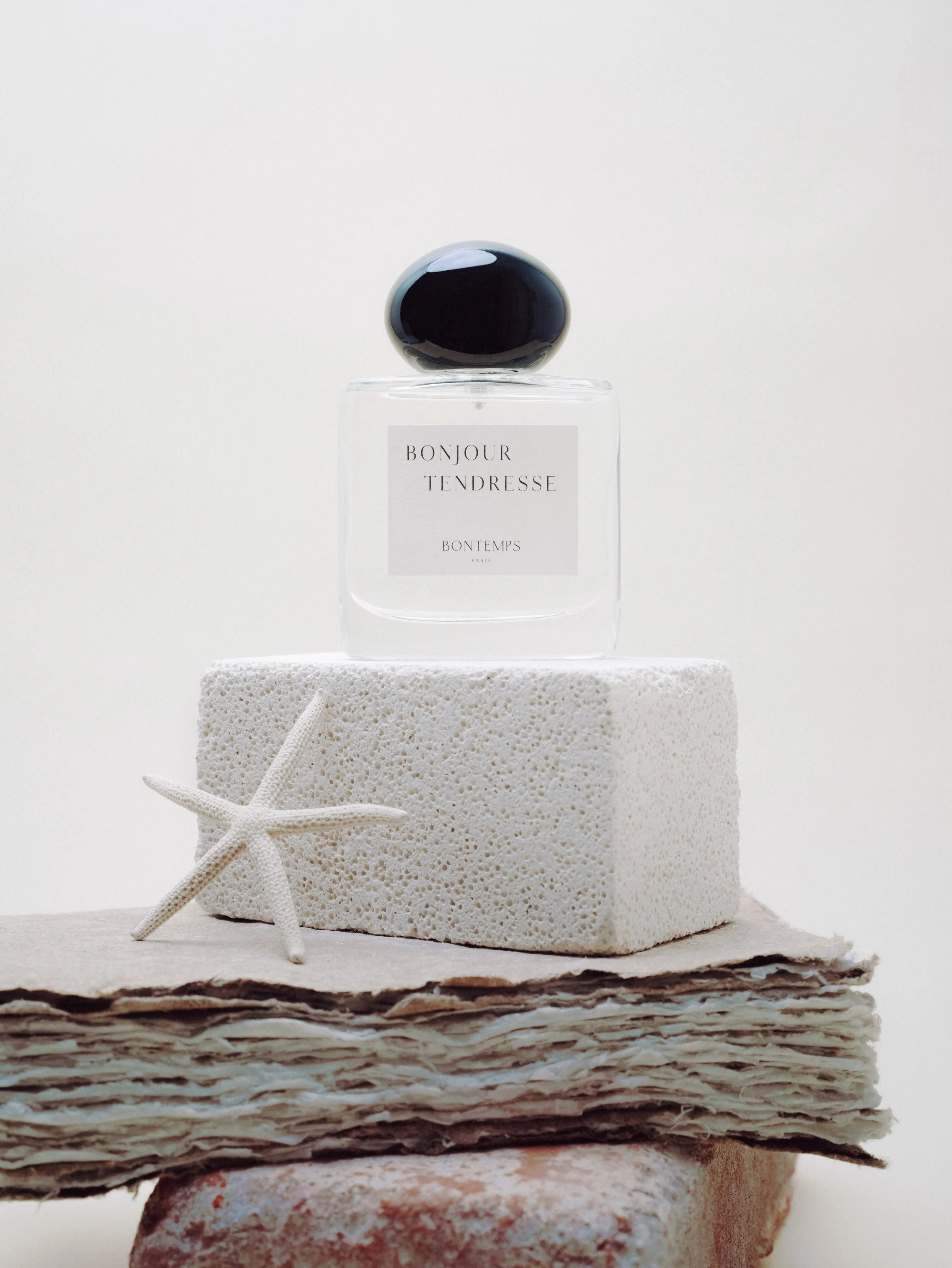 Une bouteille de parfum transparente avec un étiquette blanche, un couvercle noir brillant, posée sur une éponge blanche, avec une étoile de mer en plastique à côté, sur un tas de papiers et de pierres.