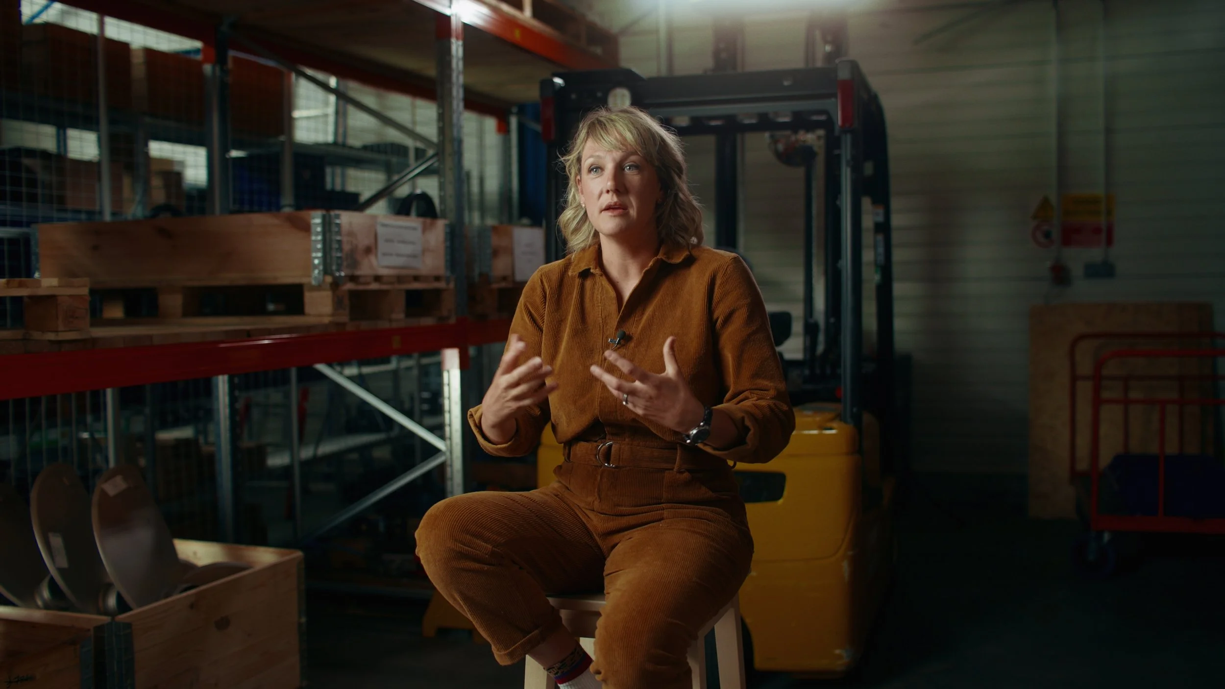 Une femme assise sur un tabouret, parlant dans un environnement d'entrepôt ou de stockage avec des étagères en métal, des palettes en bois, un chariot élévateur en arrière-plan et un mur en métal en arrière.