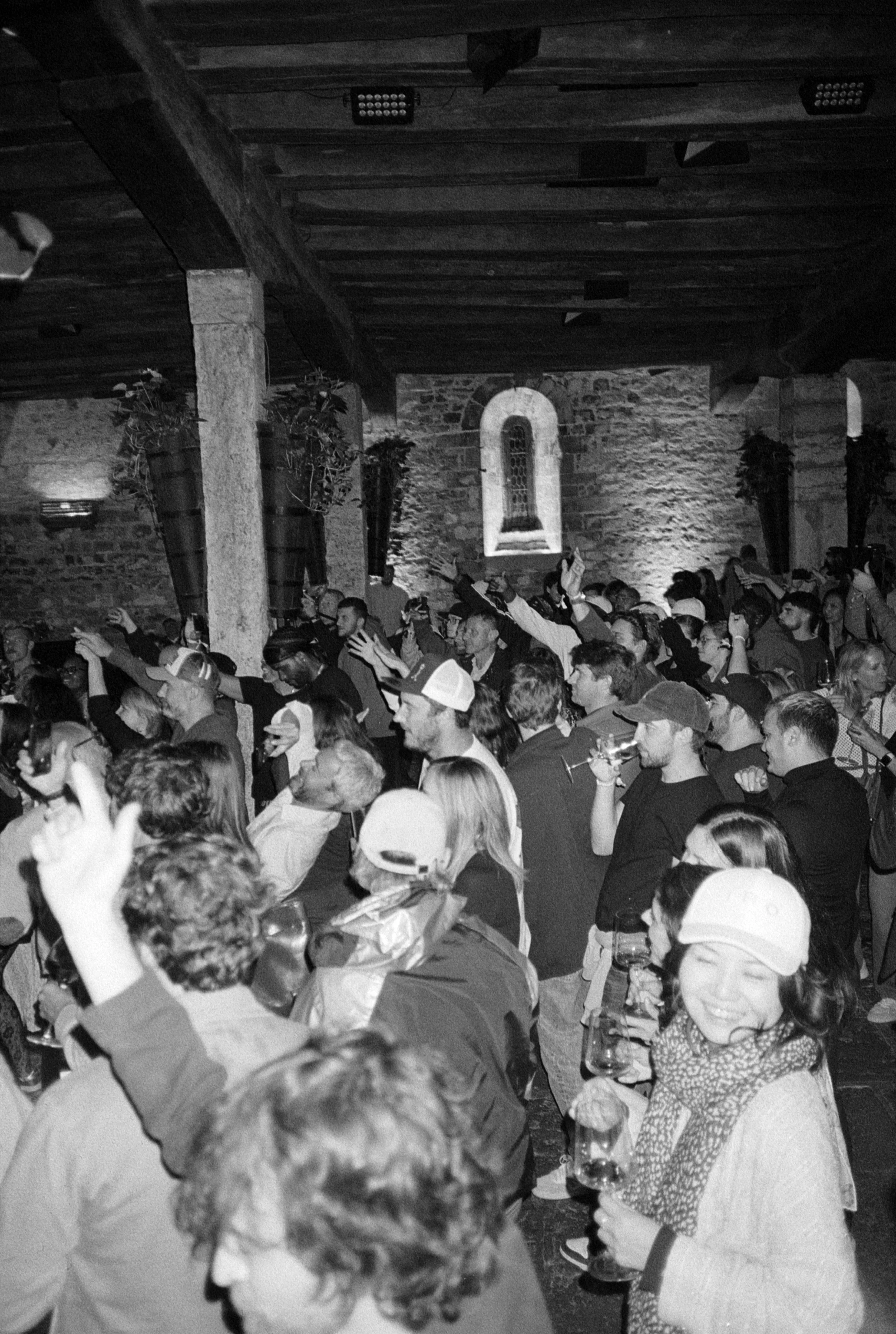 Une foule de personnes à un événement dans une salle avec murs en pierre, certains levant les mains ou tenant des verres, ambiance animée.