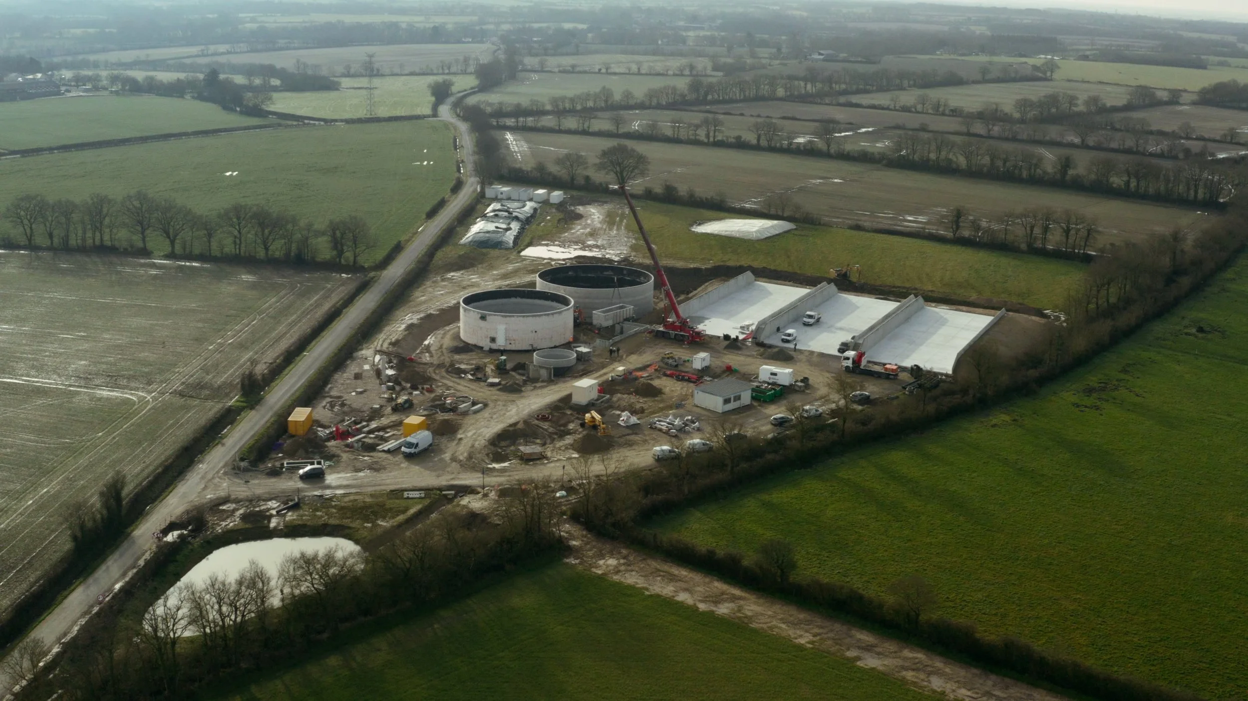Un site de construction avec deux grandes cuves circulaires et une structure de toiture en cours d'installation, entouré de véhicules et d'équipements, situé dans un cadre rural avec des champs et des arbres.