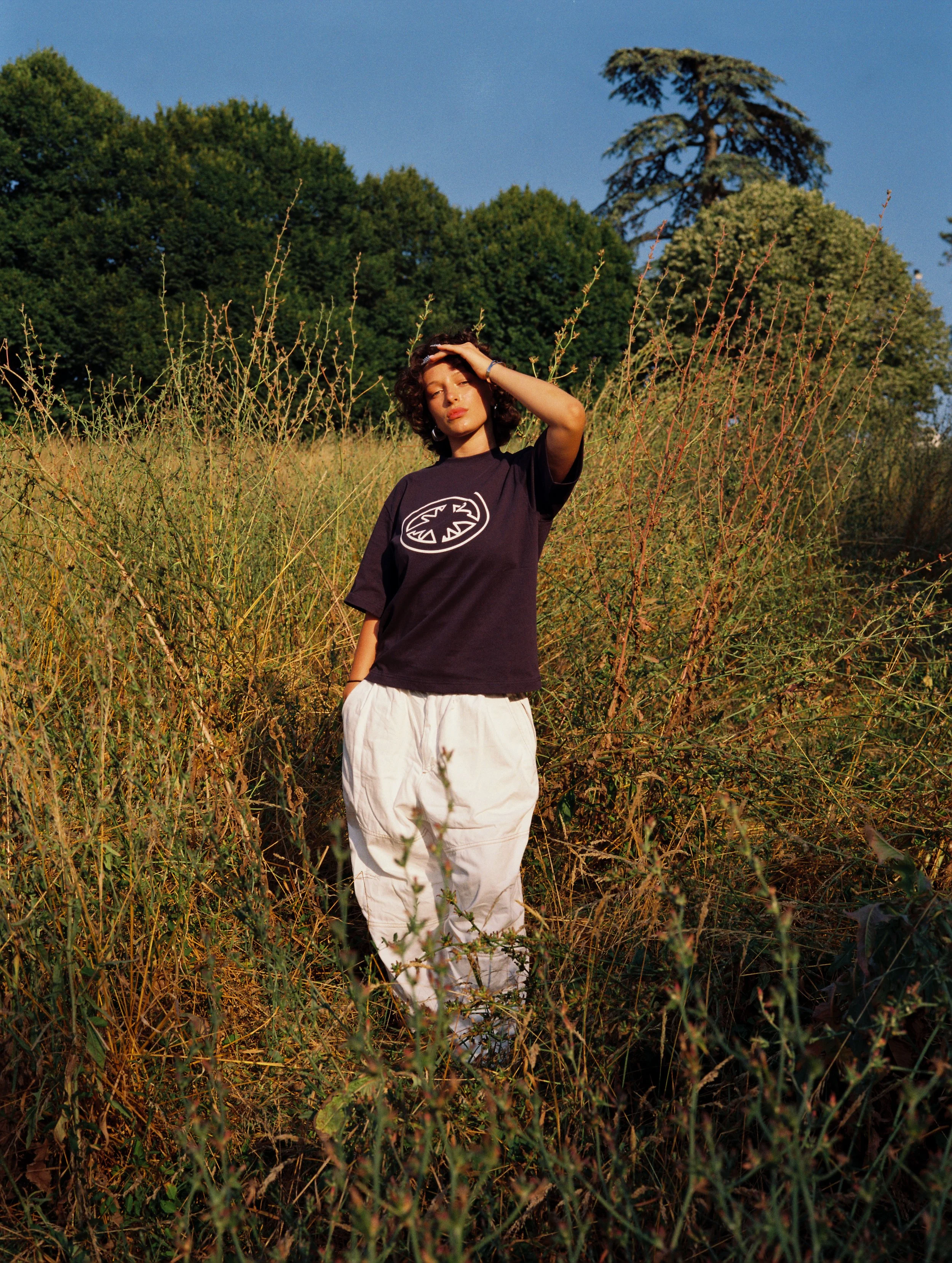Une femme debout dans un champ d'herbes hautes, portant un t-shirt noir avec un logo blanc et un pantalon blanc, sous un ciel bleu avec des arbres en arrière-plan.