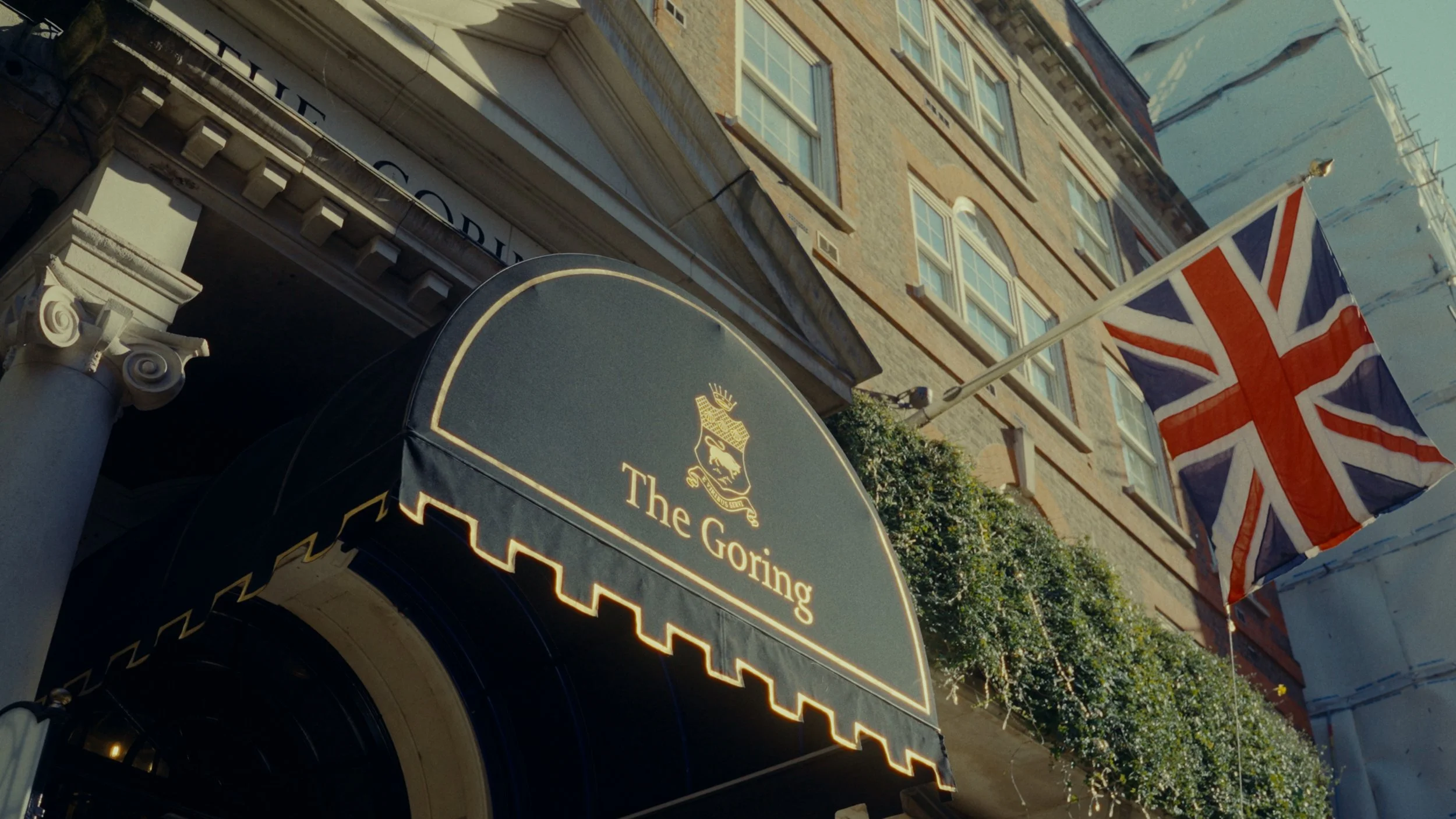 Façade d'un hôtel avec une enseigne noire portant le nom 'The Goring' et un drapeau du Royaume-Uni flottant à côté. La façade est en briques et il y a des fenêtres avec des volets blancs.