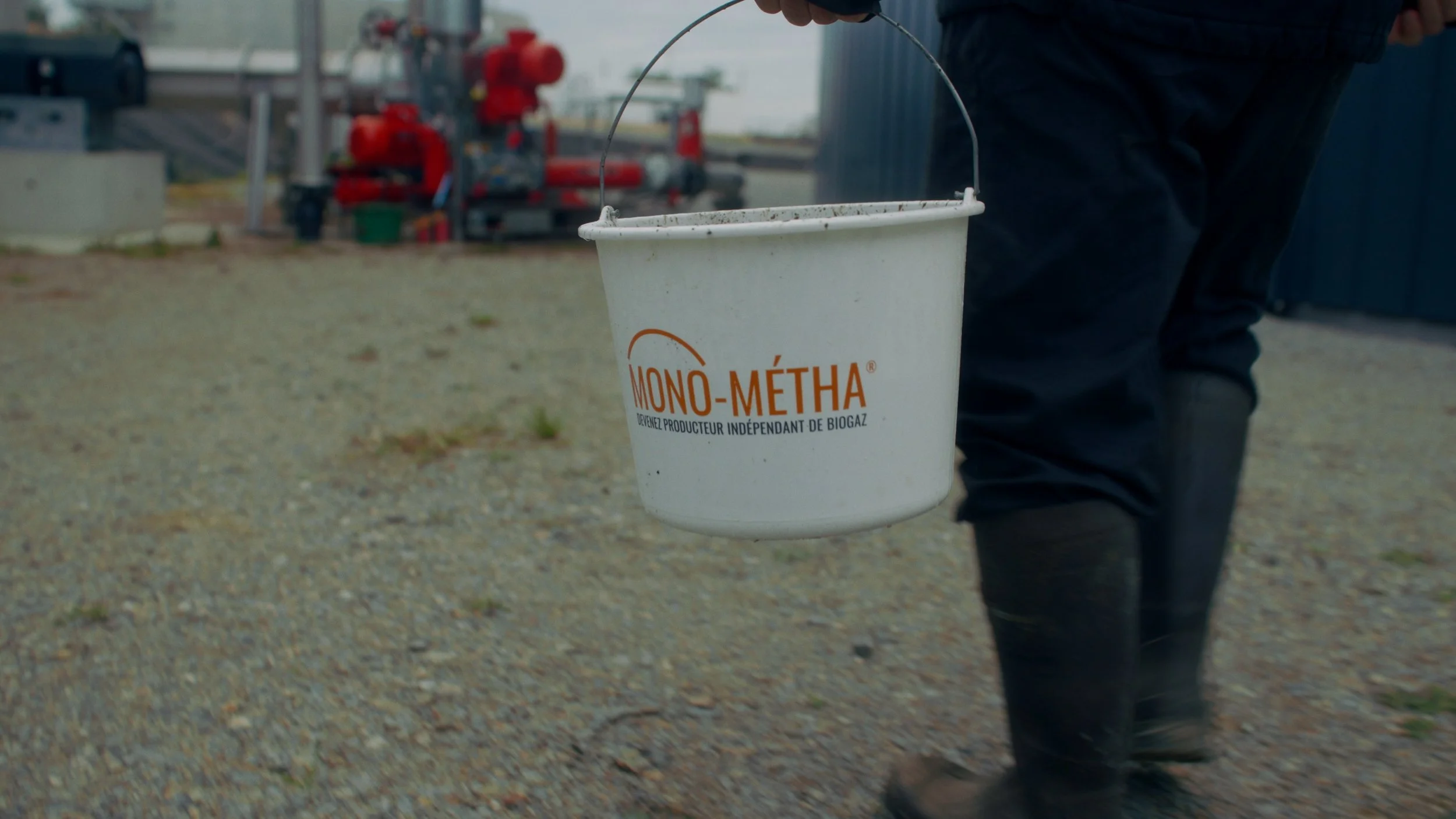 Person tenant un seau blanc avec l'inscription 'Mono-Métha' en orange, en dehors d'une usine ou d'une installation industrielle.
