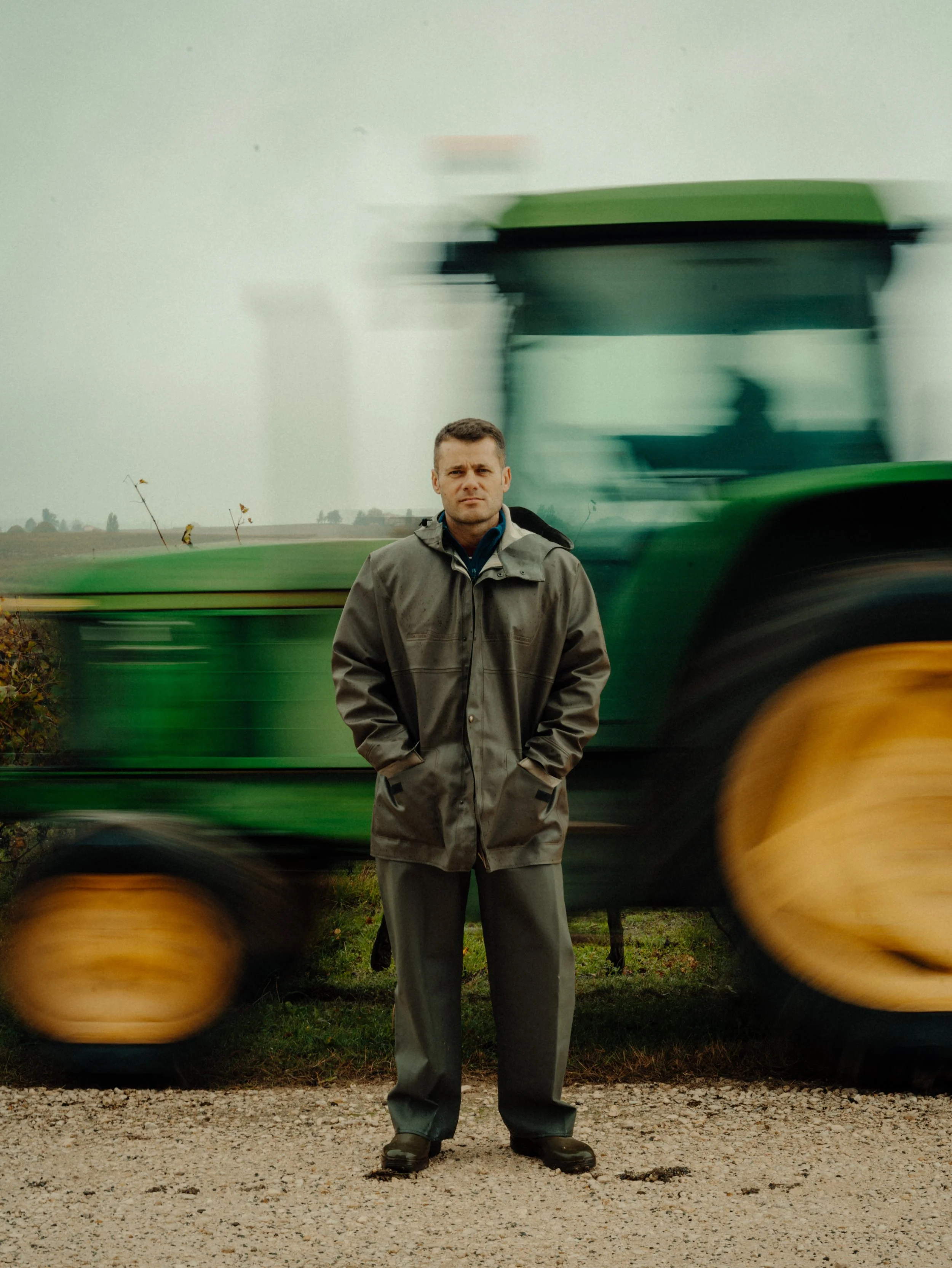 Homme en manteau gris debout devant un tracteur vert en mouvement dans un champ.