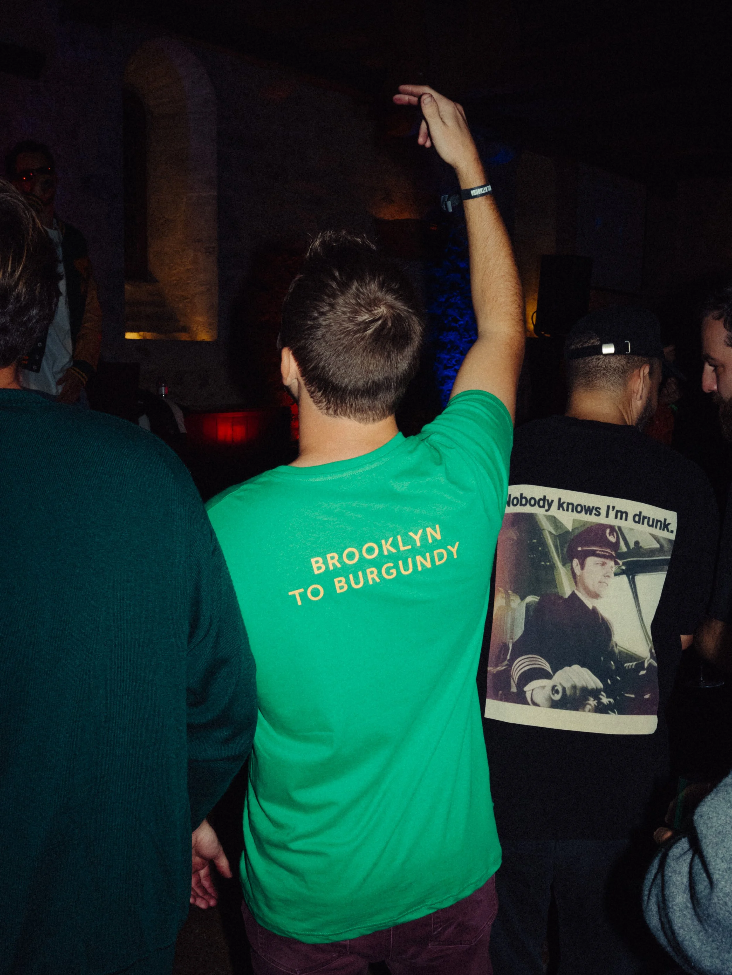 Groupe de jeunes gens dans une fête ou un événement, certains portant des t-shirts avec des textes humoristiques ou locaux, dans une ambiance sombre avec des lumières colorées.