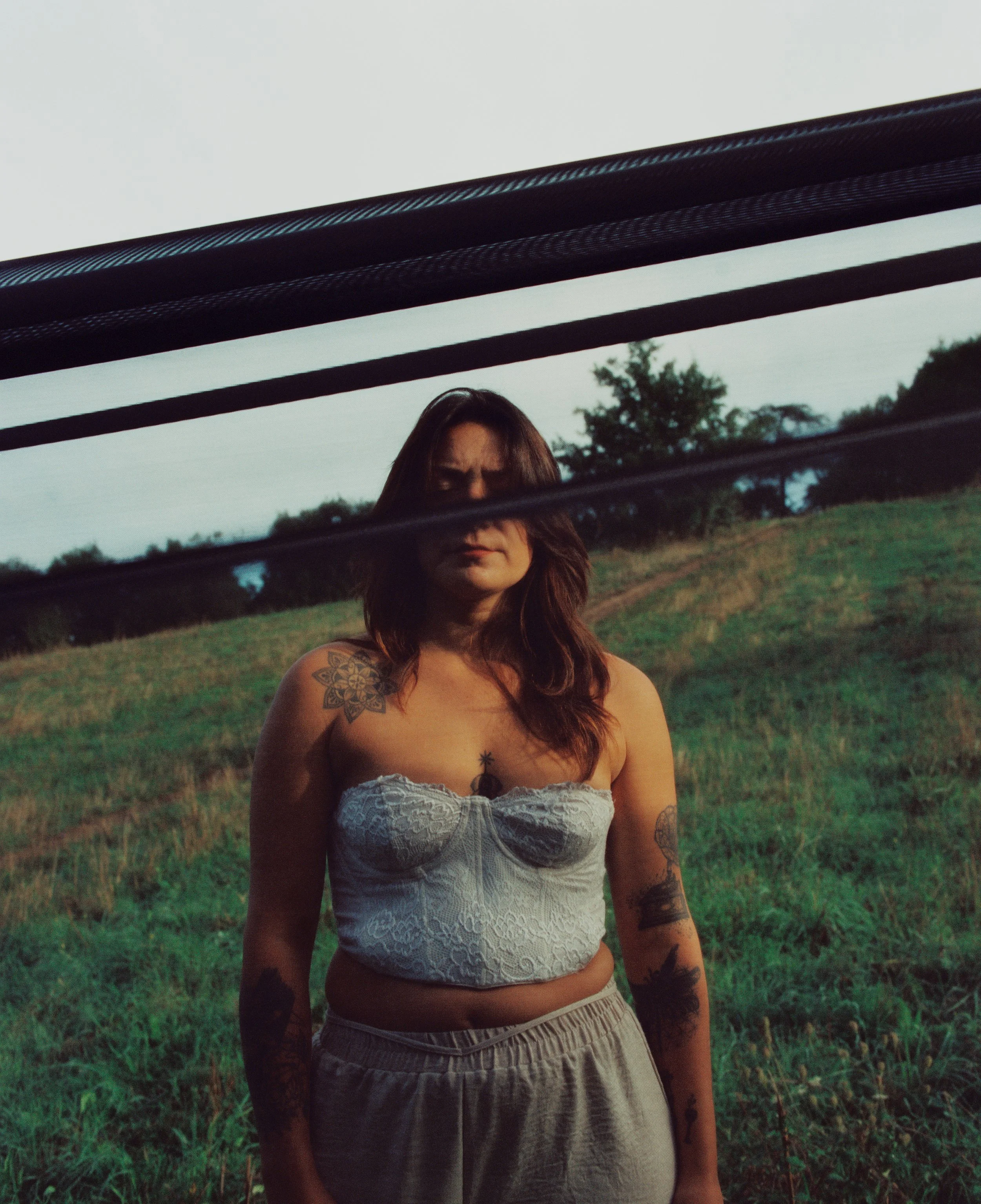 Une femme debout dans un champ vert, vue à travers une vitre avec des lignes en haut, portant un haut blanc en dentelle et des pantalons amples, avec des tatouages sur ses bras, les cheveux bruns et longs.