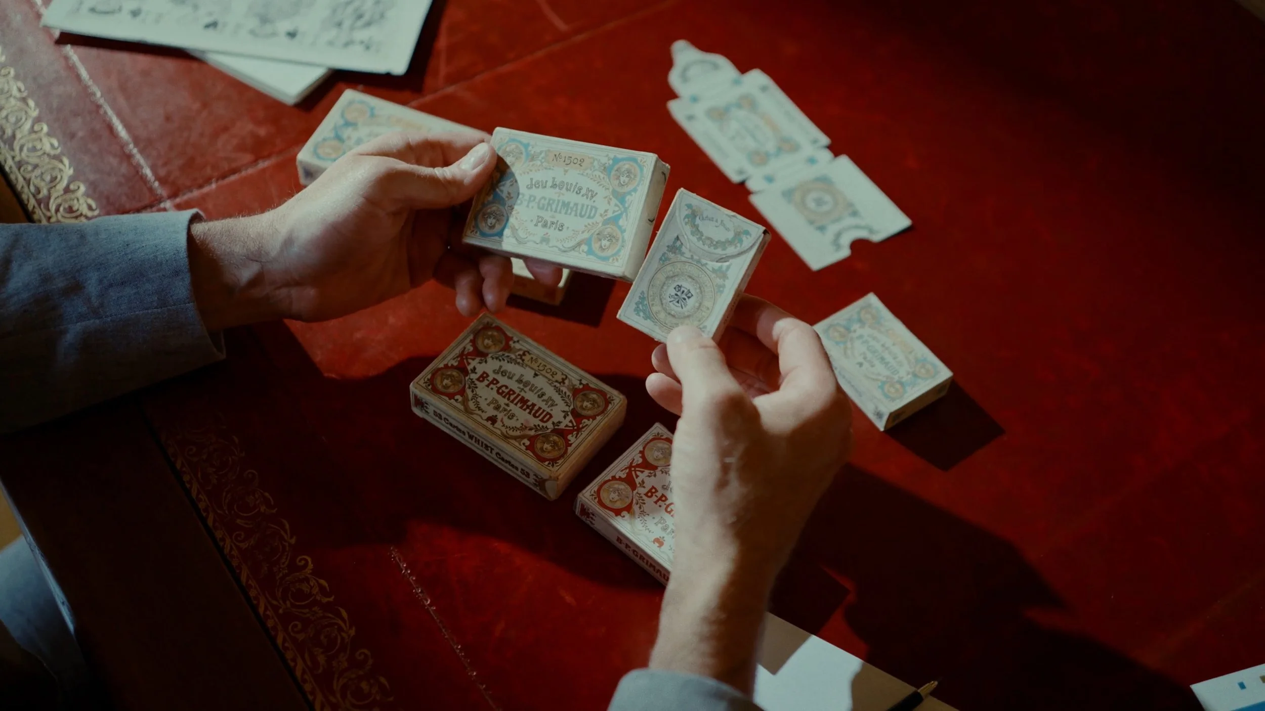Deux personnes jouent aux cartes de tarot sur une table en bois rouge. L'une d'elles tient un paquet de cartes colorées intitulé 'Jeu Louis XV B.P. Grimau'.