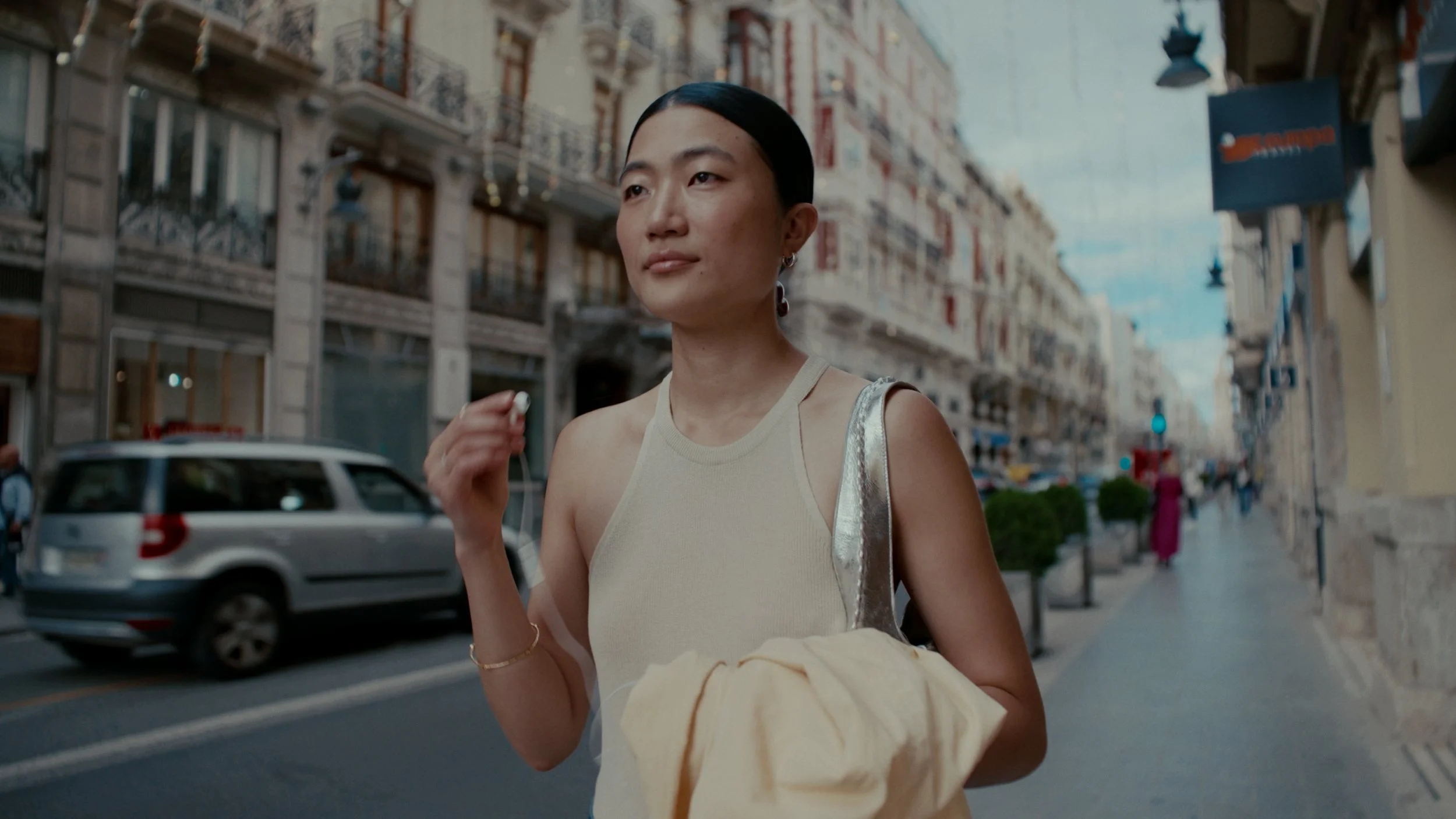 Une femme marche dans une rue urbaine, portant un sac à main argenté et tenant un vêtement beige. La scène est éclairée par la lumière naturelle, avec des bâtiments anciens en arrière-plan.