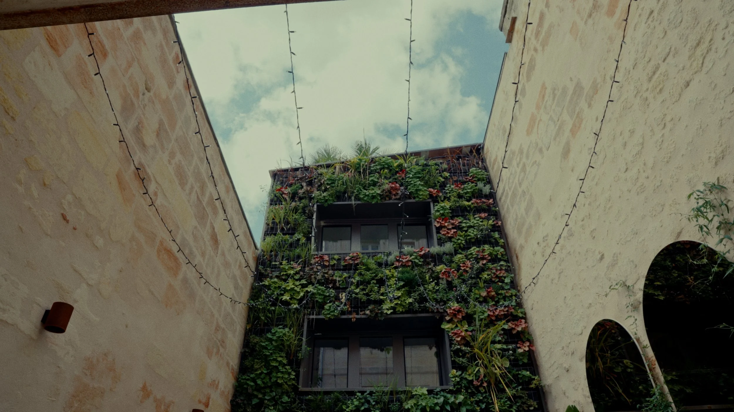 Vue intérieure d'une cour de bâtiment avec un mur végétal en façade, ciel bleu avec quelques nuages, et guirlandes lumineuses suspendues.