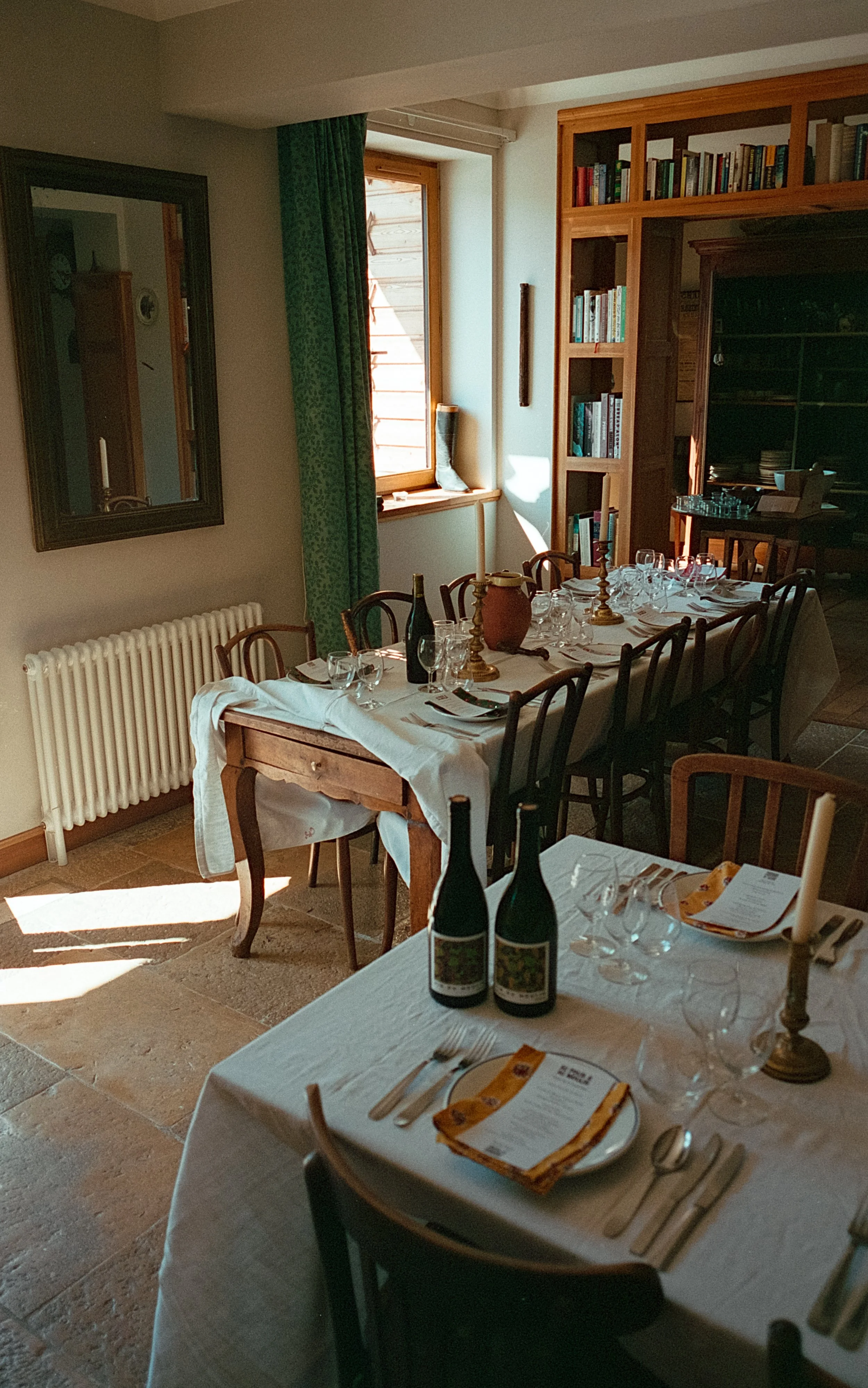 Salle à manger préparée avec une table dressée avec des bouteilles de vin, des verres, des chandeliers, des assiettes, des couverts et des menus. La pièce est éclairée par la lumière naturelle entrant par une fenêtre et il y a une bibliothèque en boi