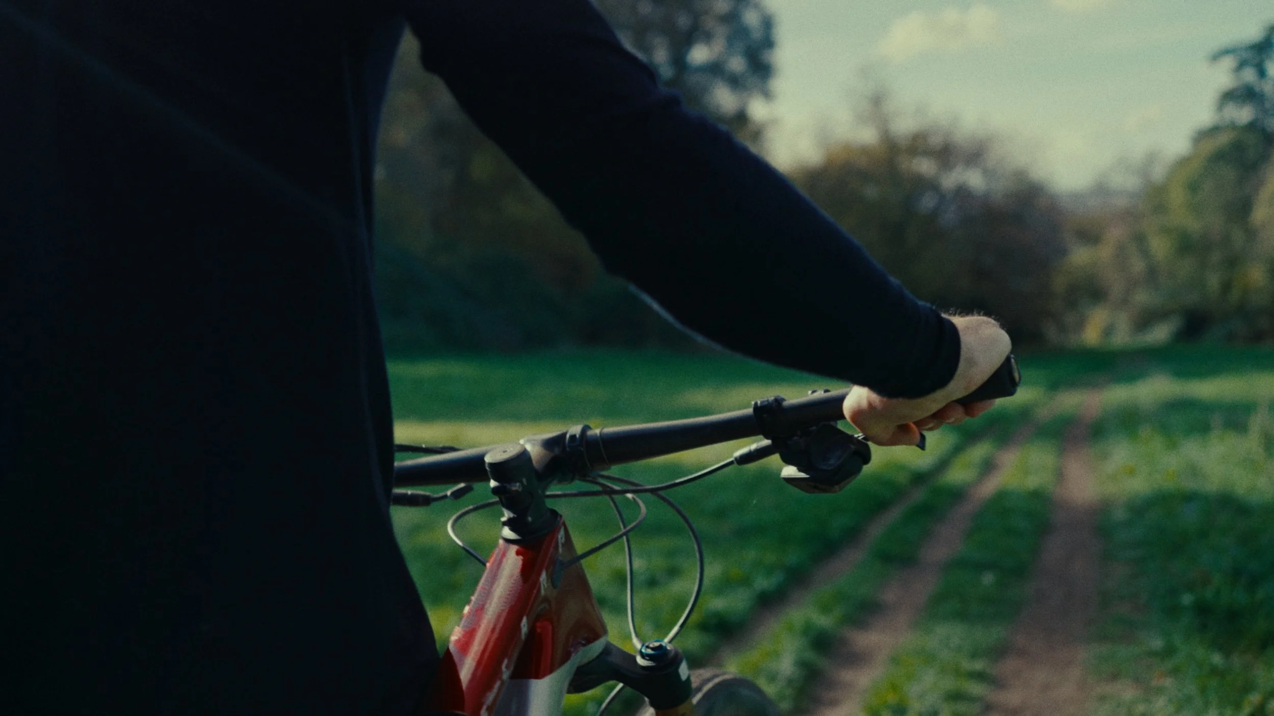 Une personne en veste noire tenant le guidon d'un vélo rouge en pleine nature, avec un chemin en terre devant, entouré d'arbres et de verdure, lors d'une journée ensoleillée.