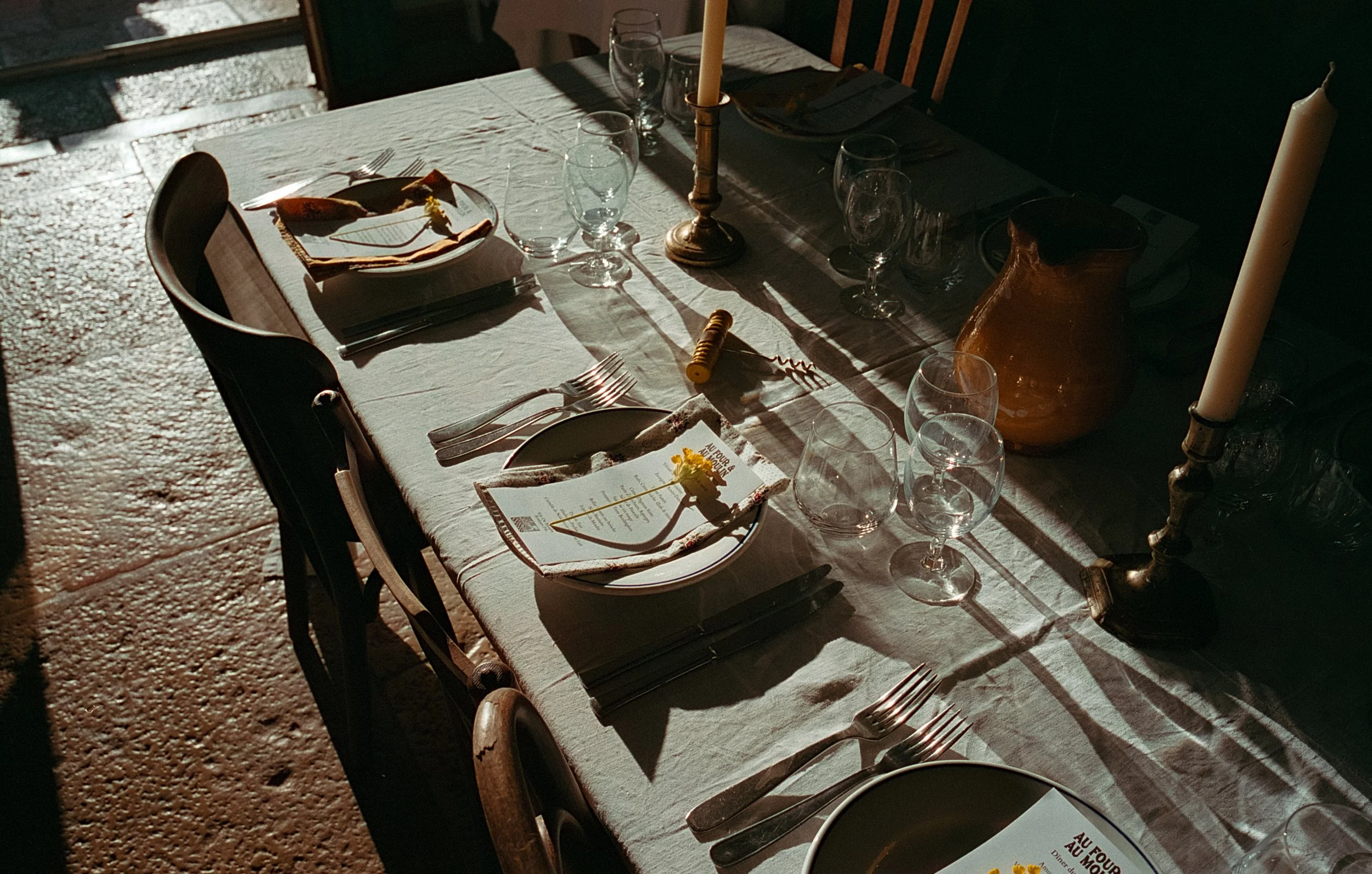 Table élégamment dressée avec une nappe blanche, des assiettes, des fourchettes, des couteaux, des verres et des bougies dans des chandeliers en argent, éclairée par la lumière du soleil.