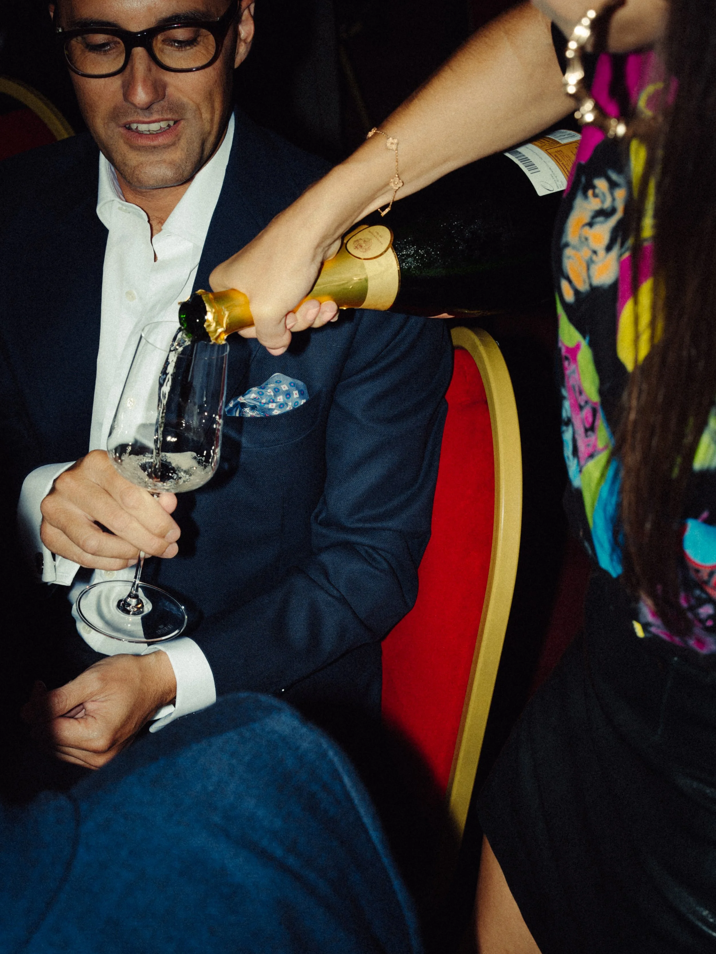 Un homme en costume noir et lunettes, assis et tenant un verre de champagne, une femme verse du champagne dans son verre, laissant couler le champagne. La scène se déroule dans un cadre intérieur.
