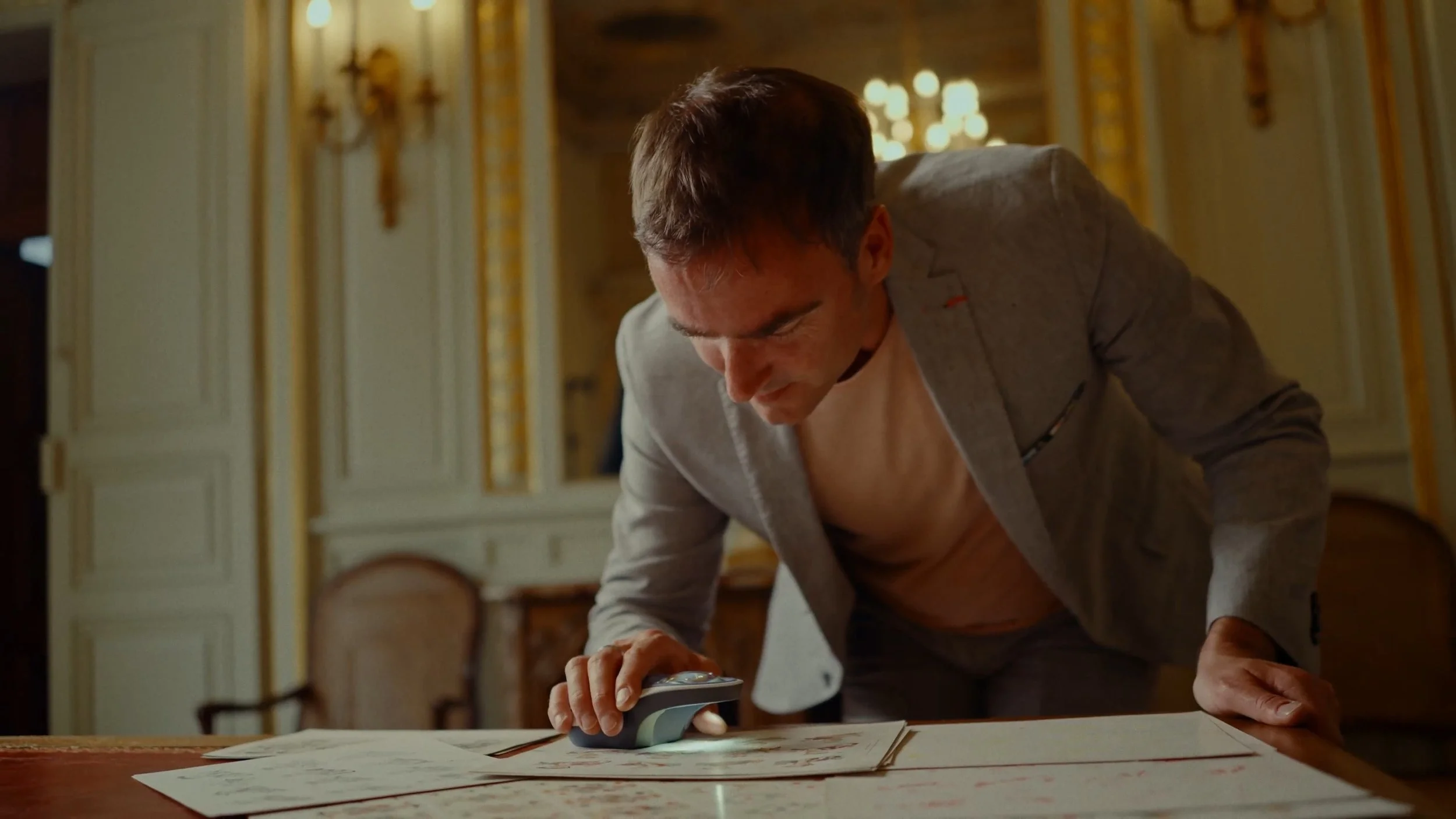 Homme en costume gris, sans chemise, qui examine des plans étendus sur une grande table dans une pièce ornée de dorures et de miroirs.