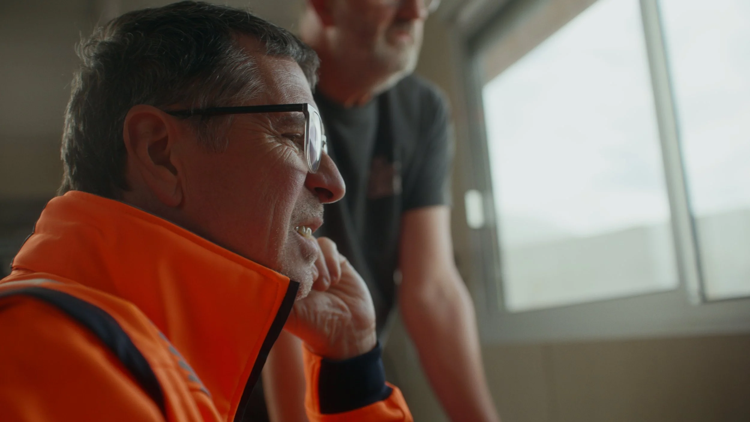 Photo de deux hommes, l'un portant des lunettes et une veste orange, apparemment en train de réfléchir ou de discuter, dans une pièce près d'une fenêtre.