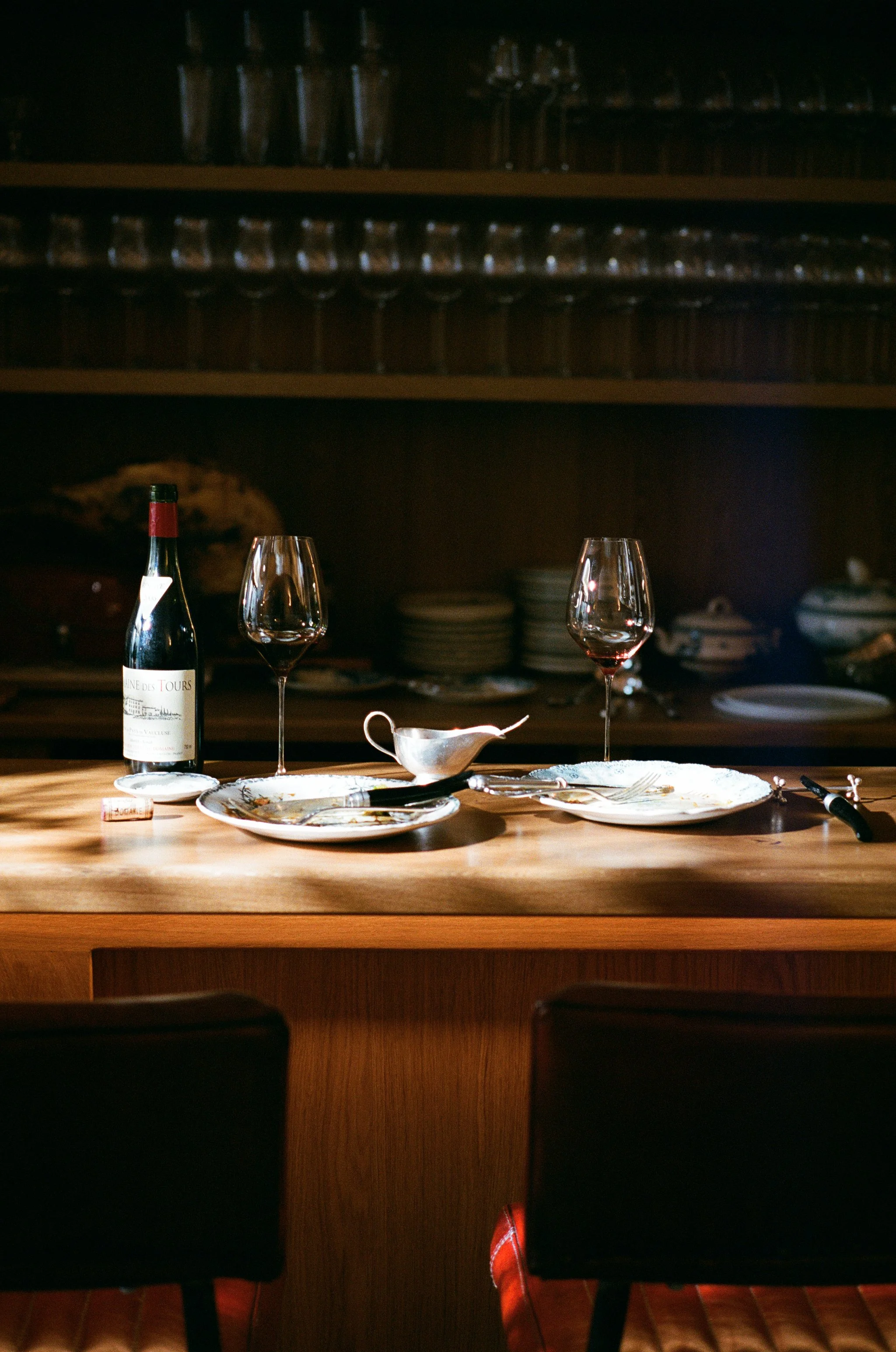 Table en bois avec deux verres à vin rouge, une bouteille de vin, une soucoupe, une petite carafe, des assiettes vides, des couverts, dans une cuisine ou un restaurant avec des étagères contenant des verres et de la vaisselle en arrière-plan.