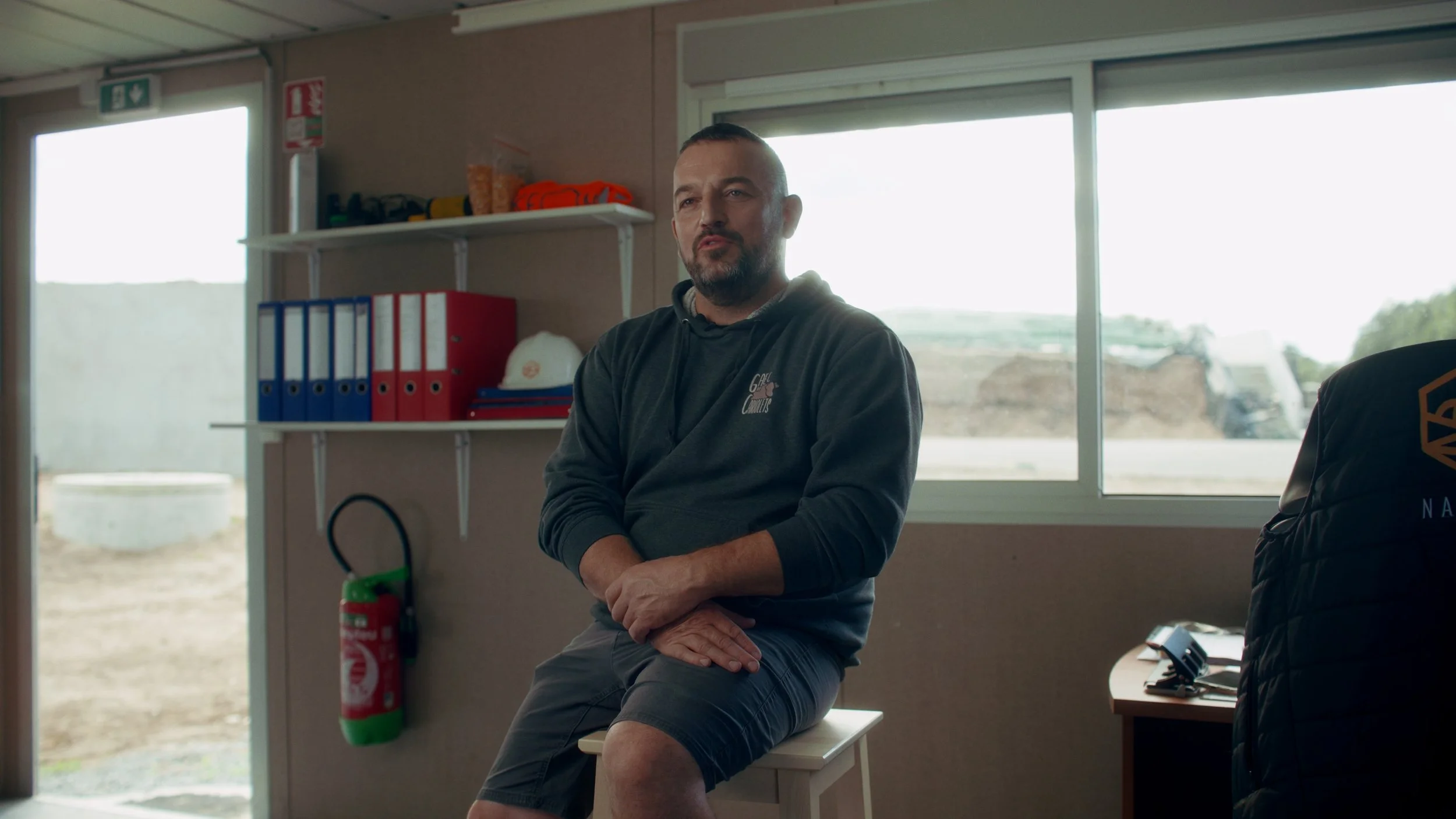 Un homme assis sur un banc dans une pièce avec une fenêtre laissant entrer la lumière. Il porte un sweat à capuche gris, des shorts gris et se trouve à côté d'une table avec un téléphone et une chaise. En arrière-plan, une étagère avec des dossiers, 