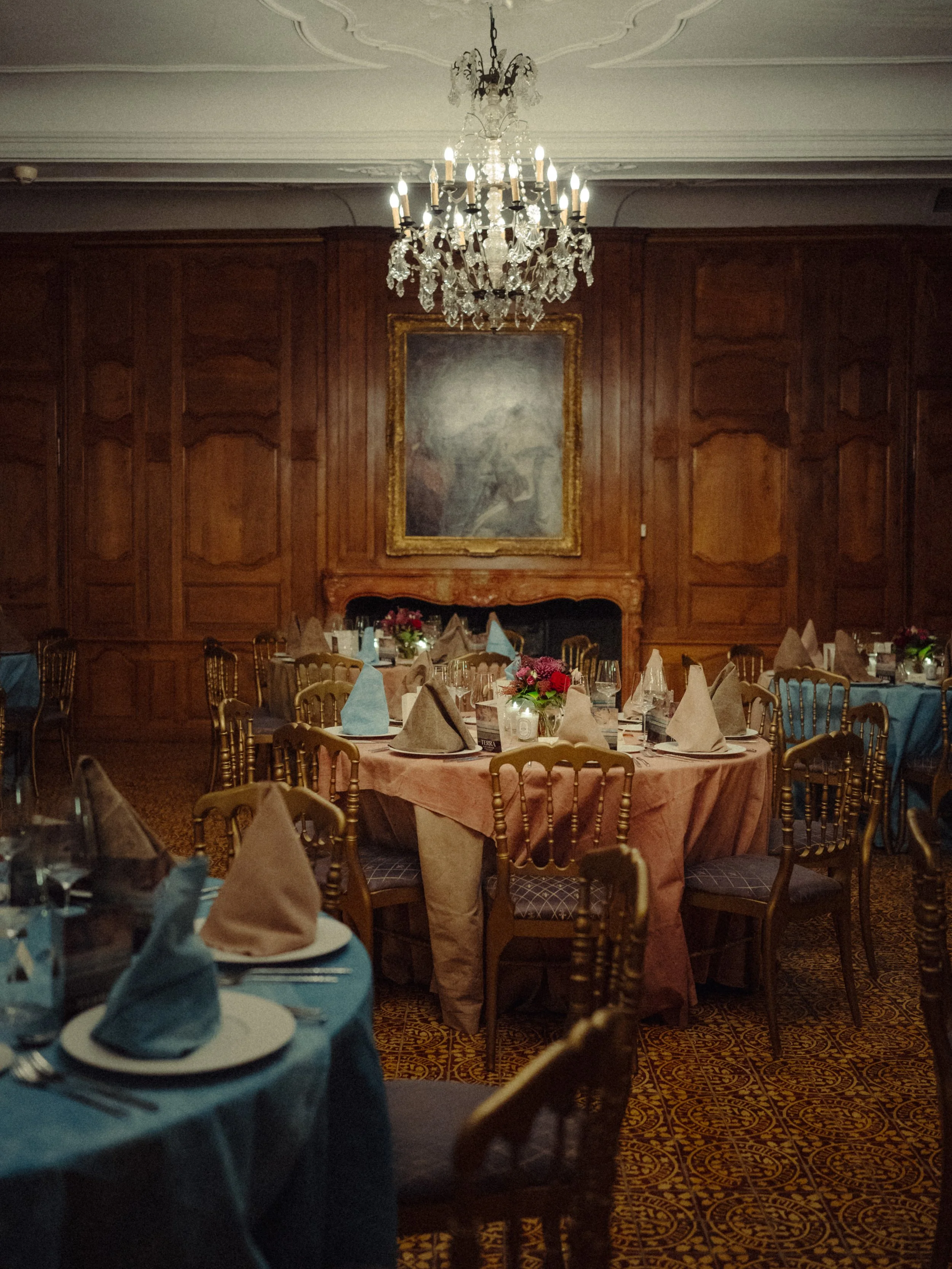 Salle de réception élégante avec tables rondes, nappes roses et bleues, serviettes pliées en pyramide, chandeliers en cristal suspendus, peinture vintage au mur, et décoration florale.