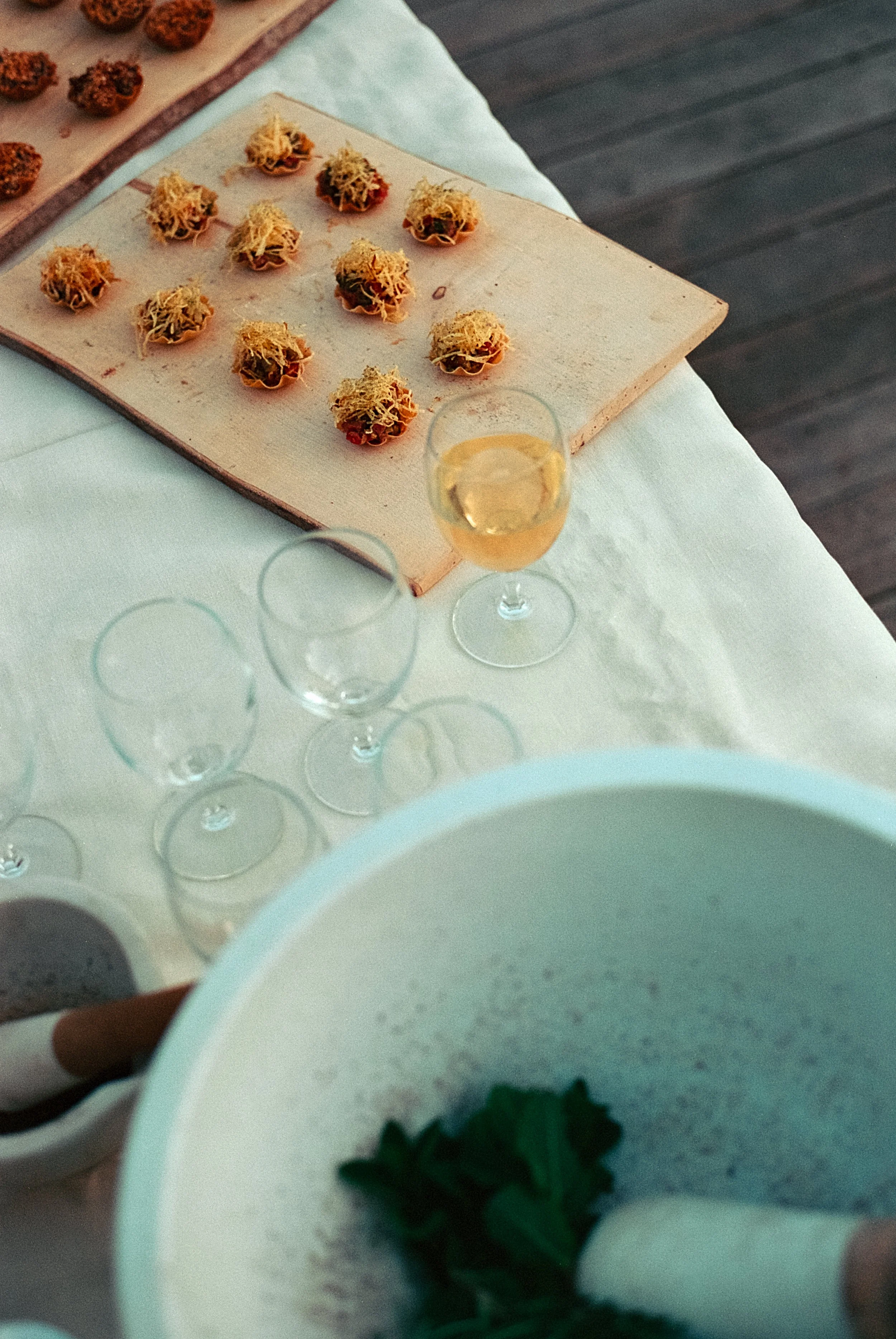 Mini tacos décoratifs sur une planche en bois, verre de boisson rosée sur une table recouverte d'une nappe blanche, un mortier avec des herbes et un pilon en premier plan.
