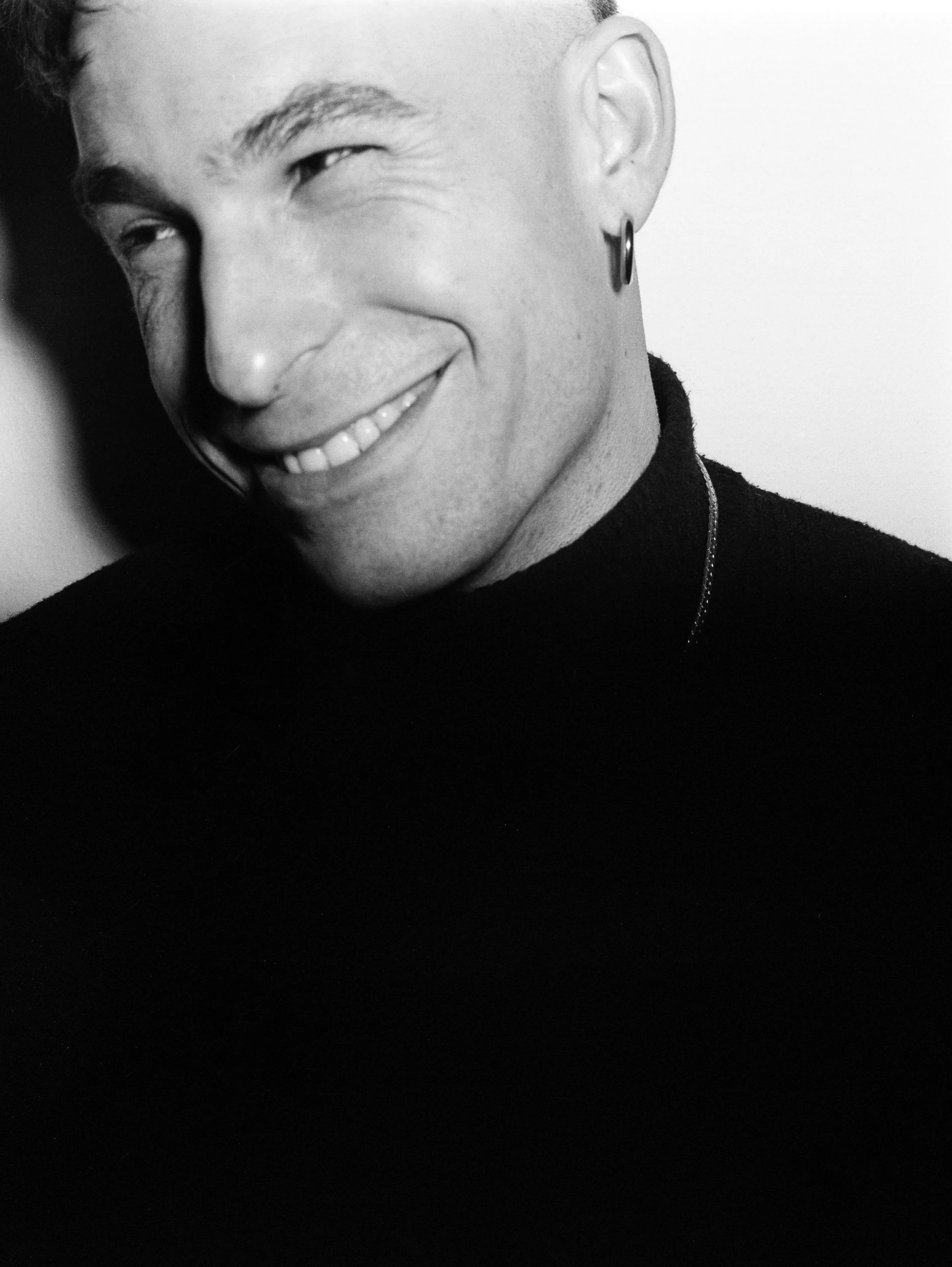 Portrait en noir et blanc d'un homme souriant, portant un piercing à l'oreille et un t-shirt noir.