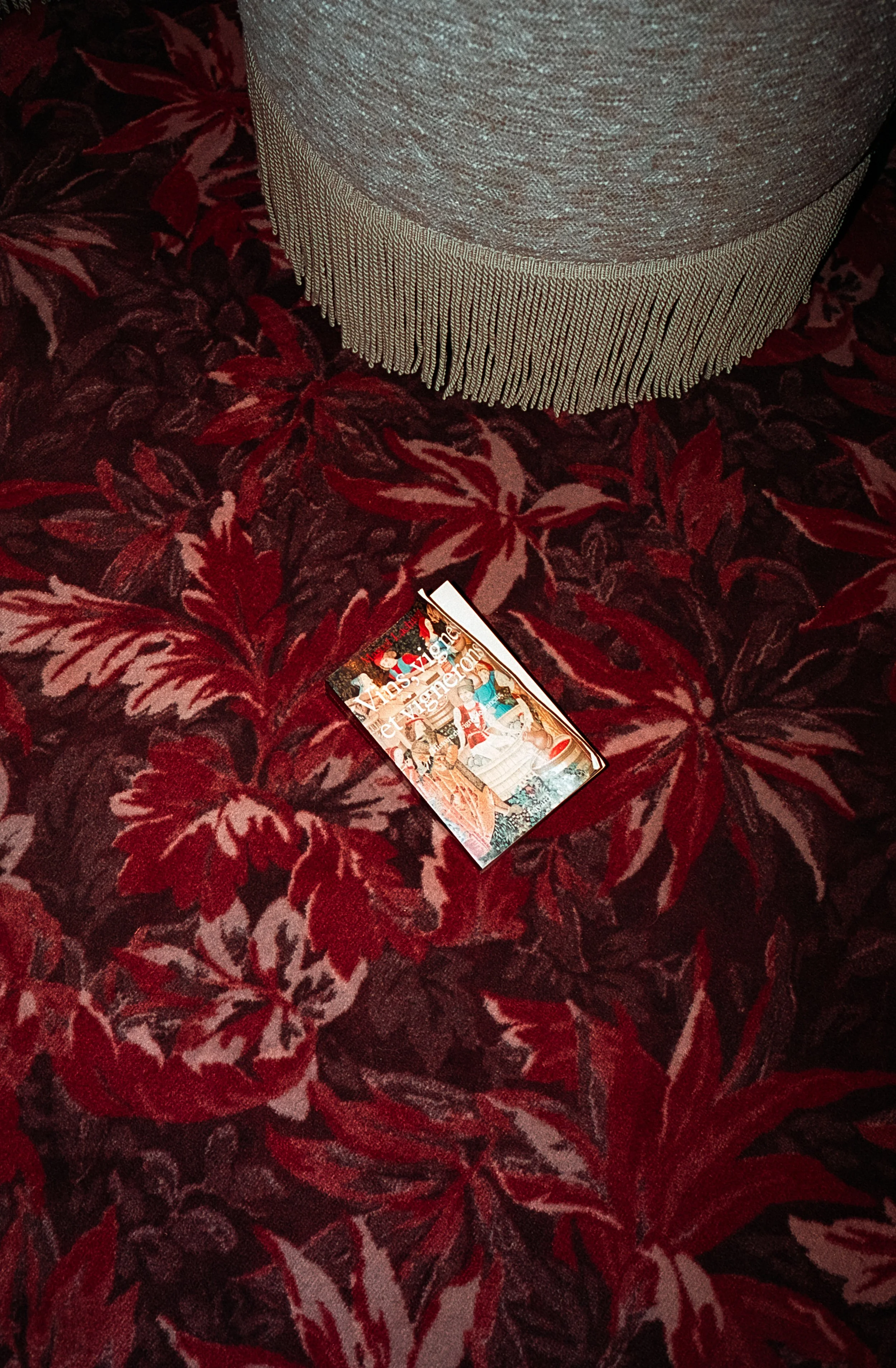 Une carte de visite ou un flyer posé sur un tapis à motif fleuri de couleur rouge et rose, avec une pièce de mobilier en tissu gris clair avec des franges en haut de l'image.