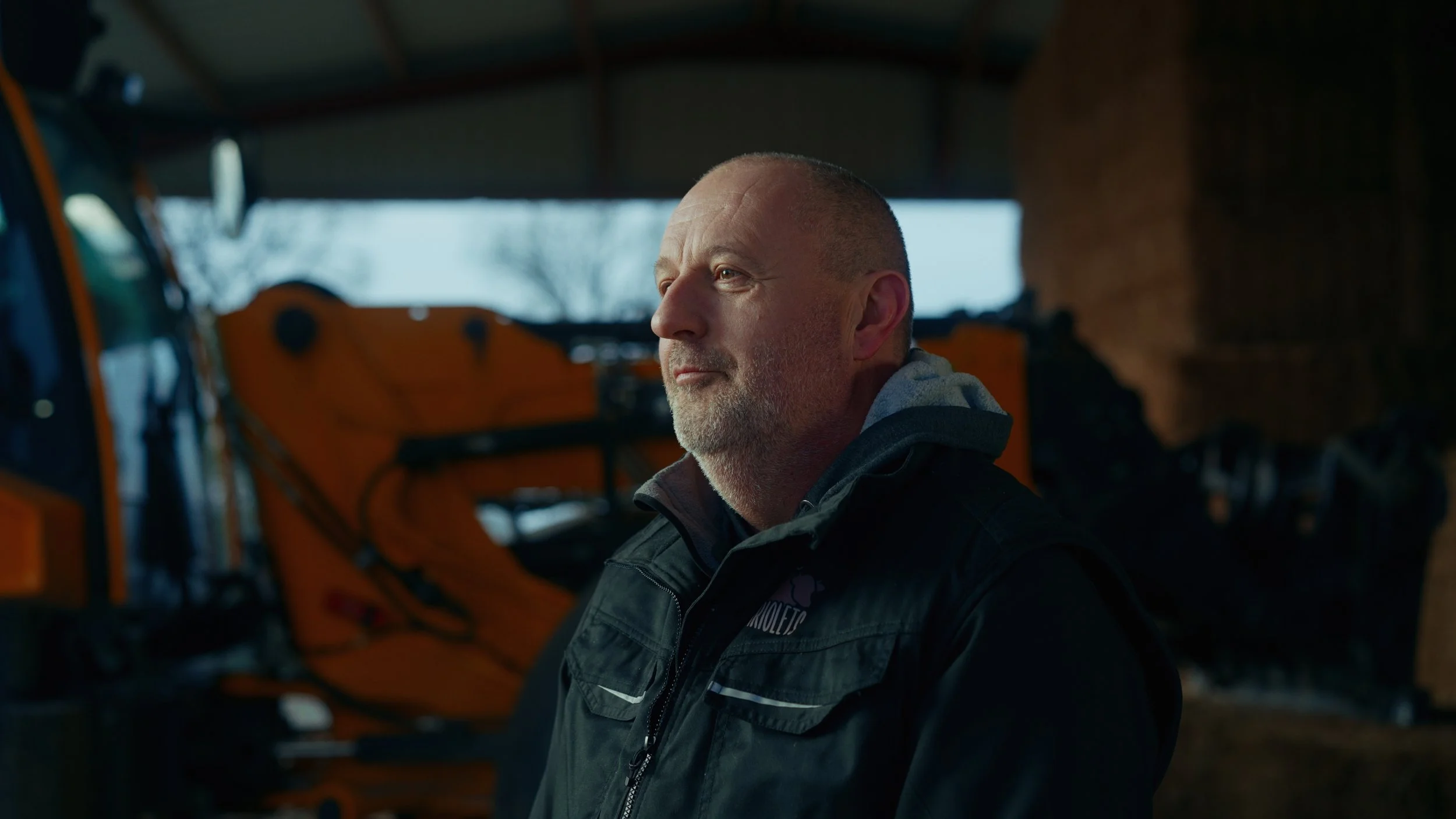 Un homme d'âge moyen avec une barbe grise, portant une veste noire, se tient dans une ferme en bois avec des machines agricoles en arrière-plan.