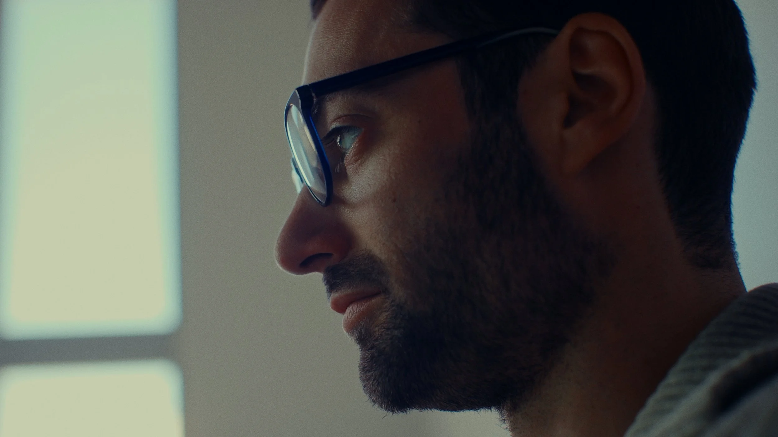 Plan rapproché d'un homme avec lunettes, regardant vers la gauche, visage du profil, lumière naturelle venant d'une fenêtre à l'arrière-plan.