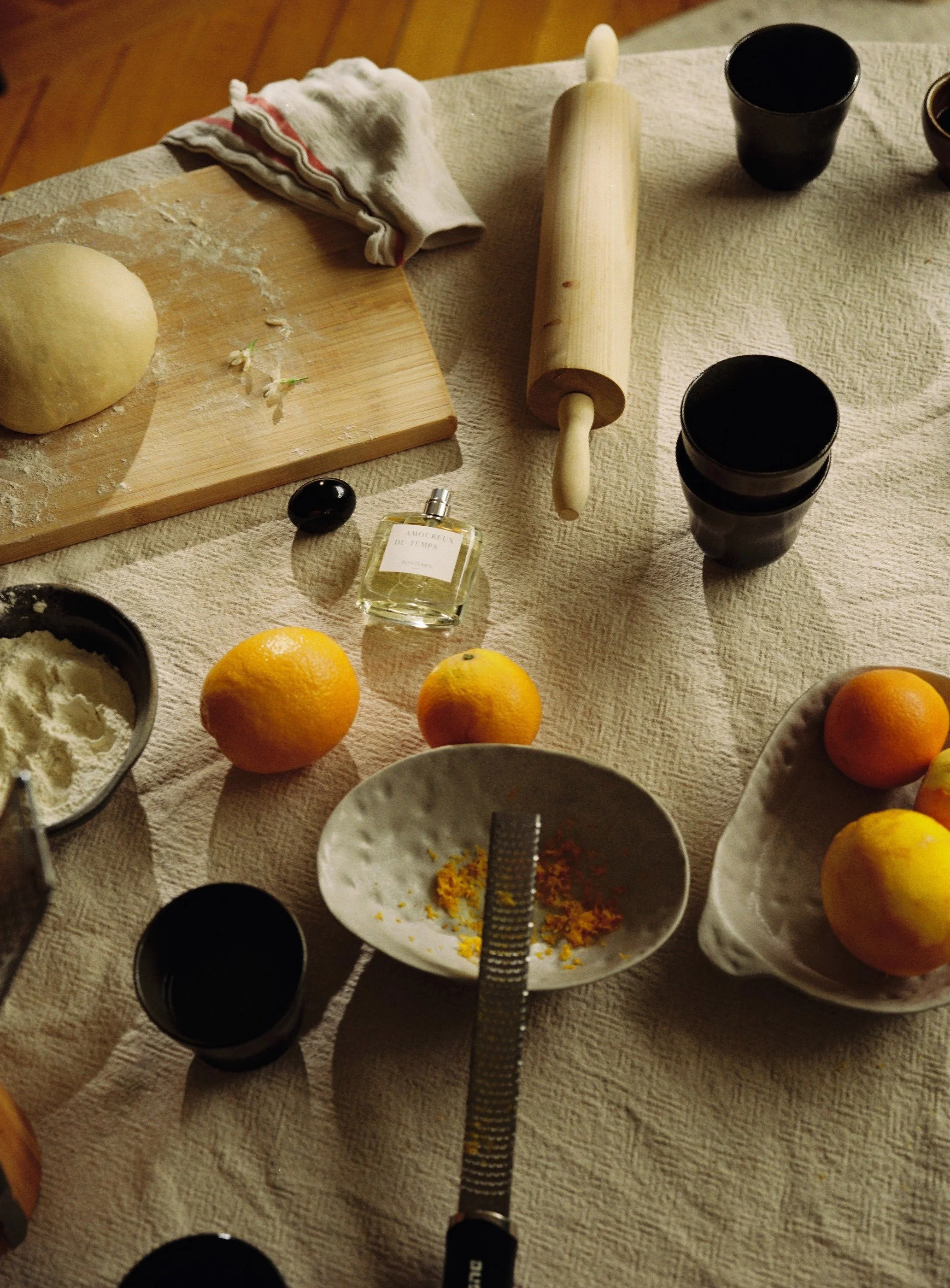 Une table de cuisine avec de la pâte, un rouleau à pâtisserie, des fruits comme des oranges et des pommes, un verre de parfum, un bol de farine, une râpe, et des tasses noires.